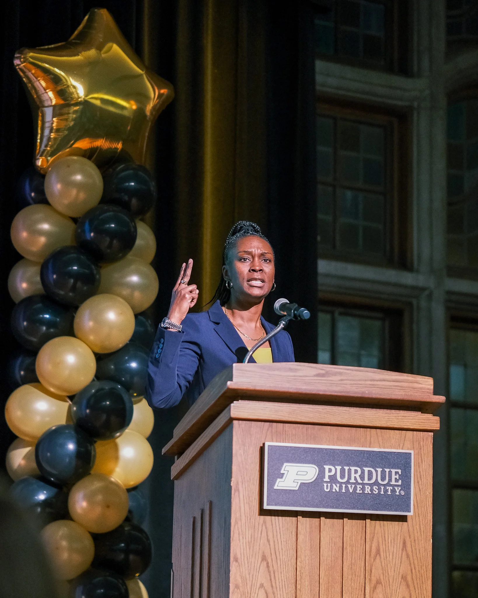 A woman speaking at a podium with a Purdue University sign, gesturing with her right hand, behind a balloon arrangement of black and gold balloons with a gold star-shaped balloon.