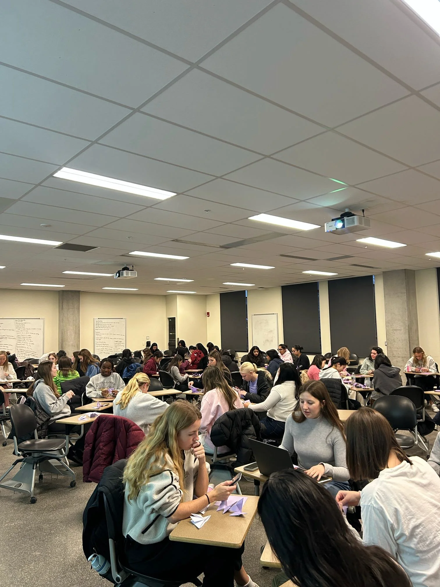 A classroom filled with students working at desks, some using laptops and others folding origami paper.