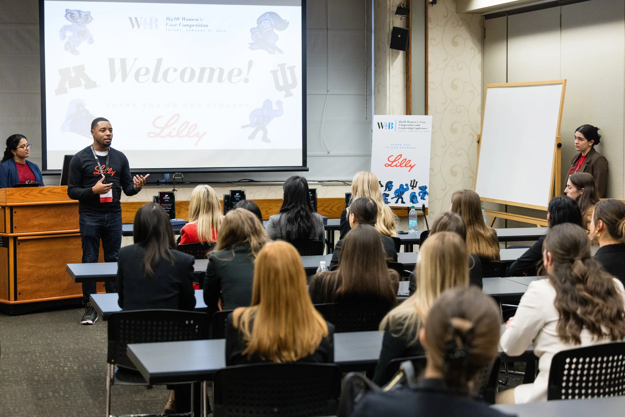 A man is giving a presentation in a conference room filled with seated women, with a large screen behind him displaying the word 'Welcome!' and a whiteboard to his right, during the Big Ten Women's Case Competition event.