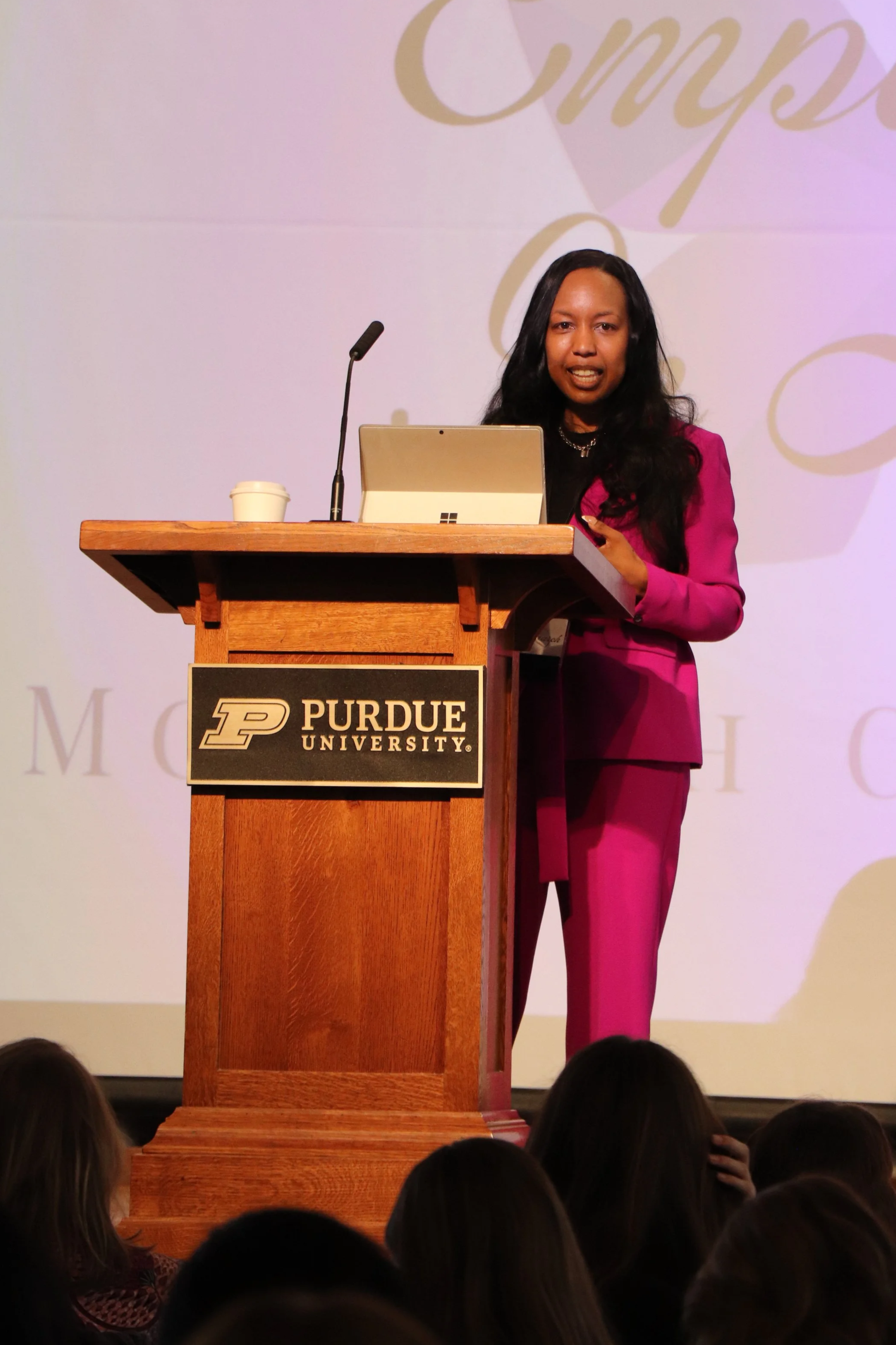 Woman in pink suit speaking at a podium with Purdue University logo, in front of a projected background.