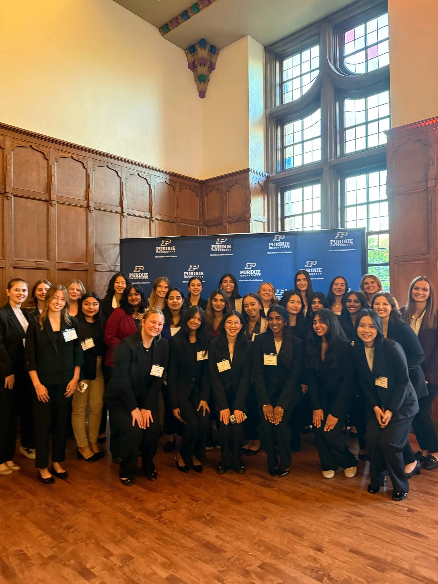 A group of young women in formal attire posing in front of a Purdue University backdrop in a large room with high windows and wood-paneled walls.