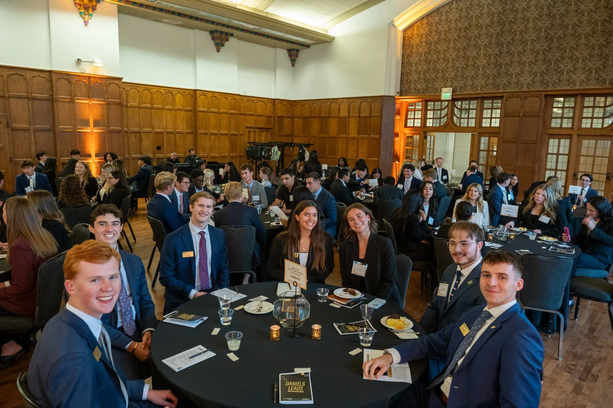 Young professionals and students attending a formal networking event or conference in a large, well-lit room with wooden paneling and high ceilings, seated around round tables with table settings and conference materials.