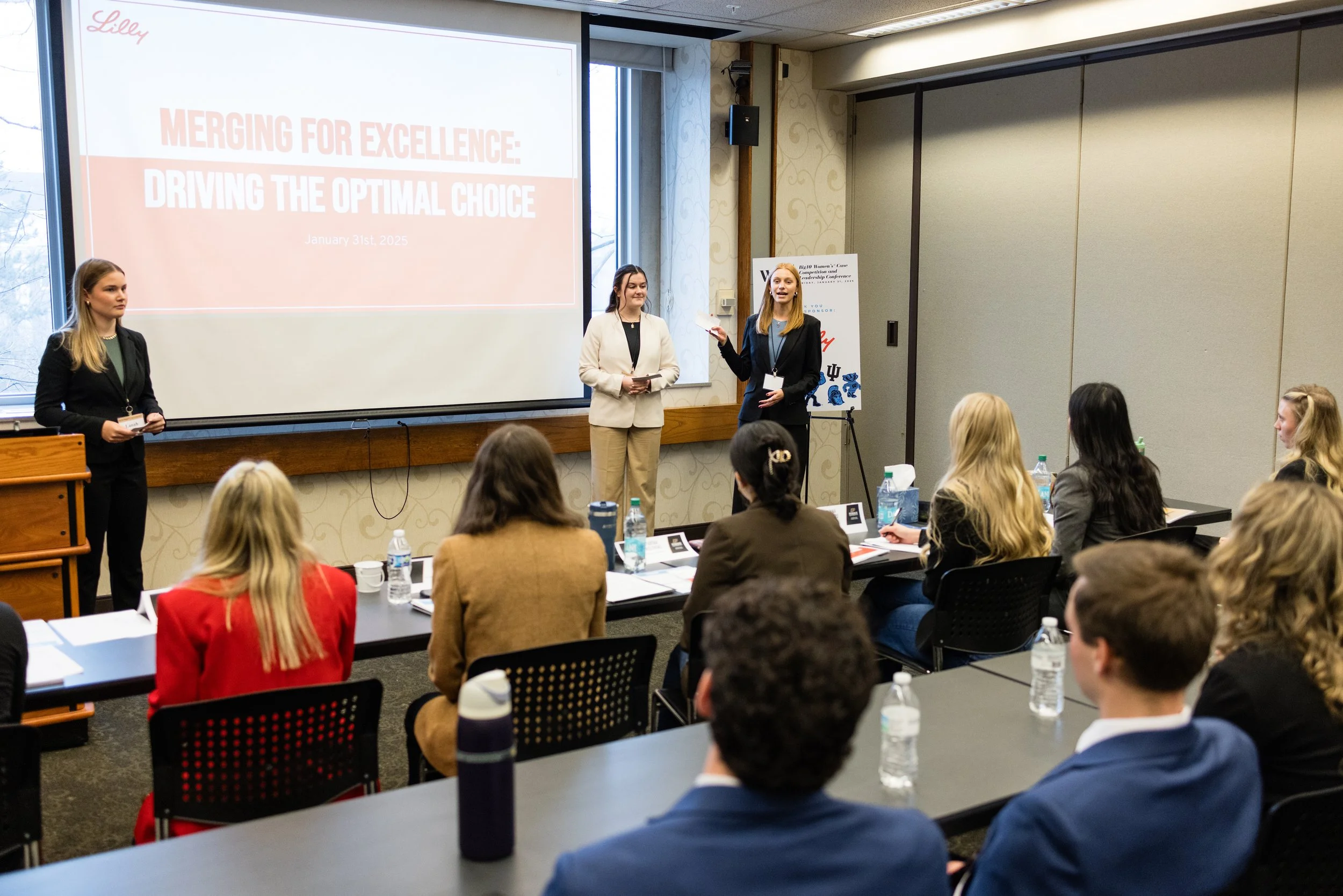 Women give a presentation to an audience in a conference room with a large screen showing a slide titled "MERGING FOR EXCELLENCE: DRIVING THE OPTIMAL CHOICE". The audience members are seated at tables with water bottles and notebooks.