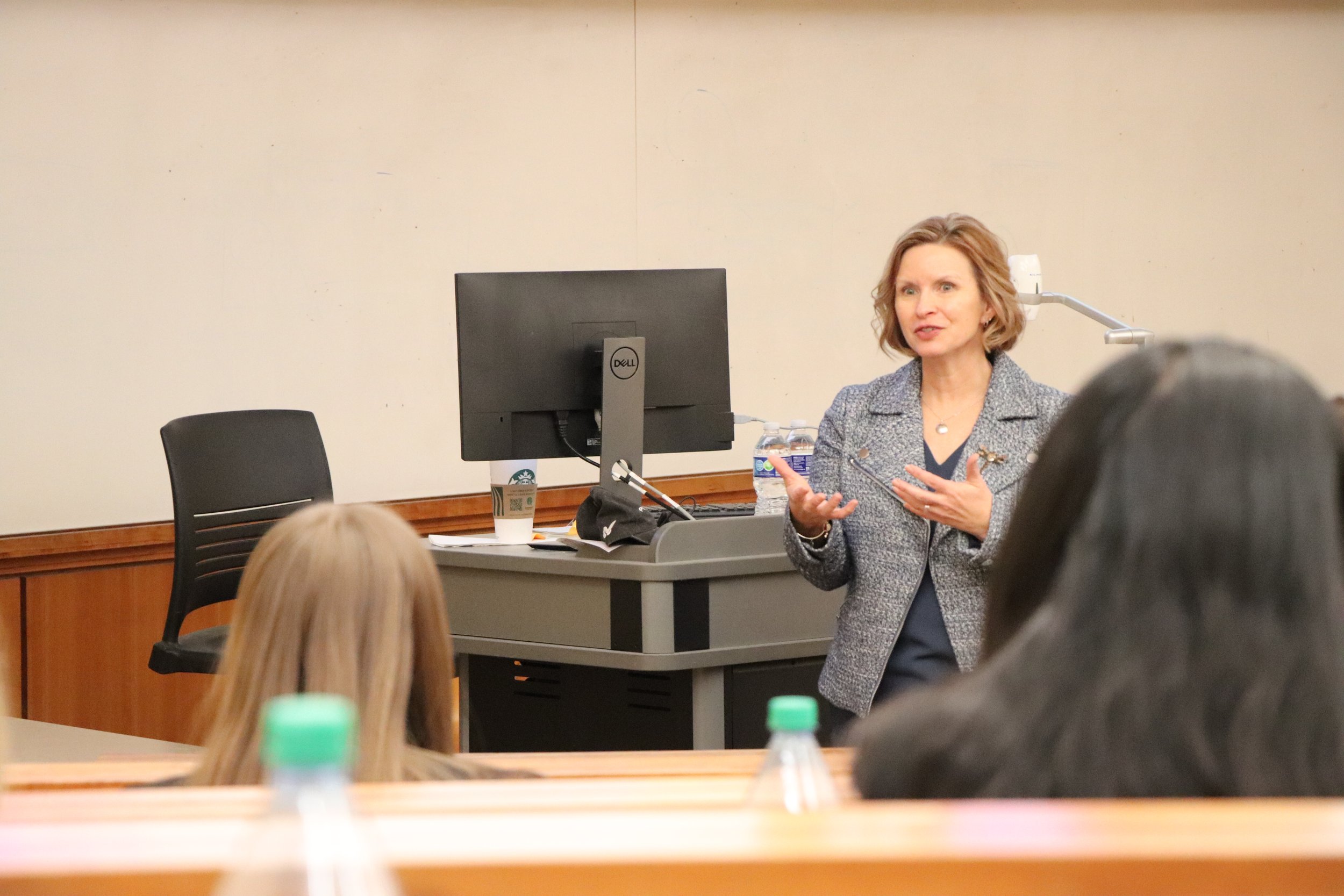 A woman with short, curly hair speaking in front of a classroom, gesturing with her hands, with a computer and water bottles on her desk, and students listening.
