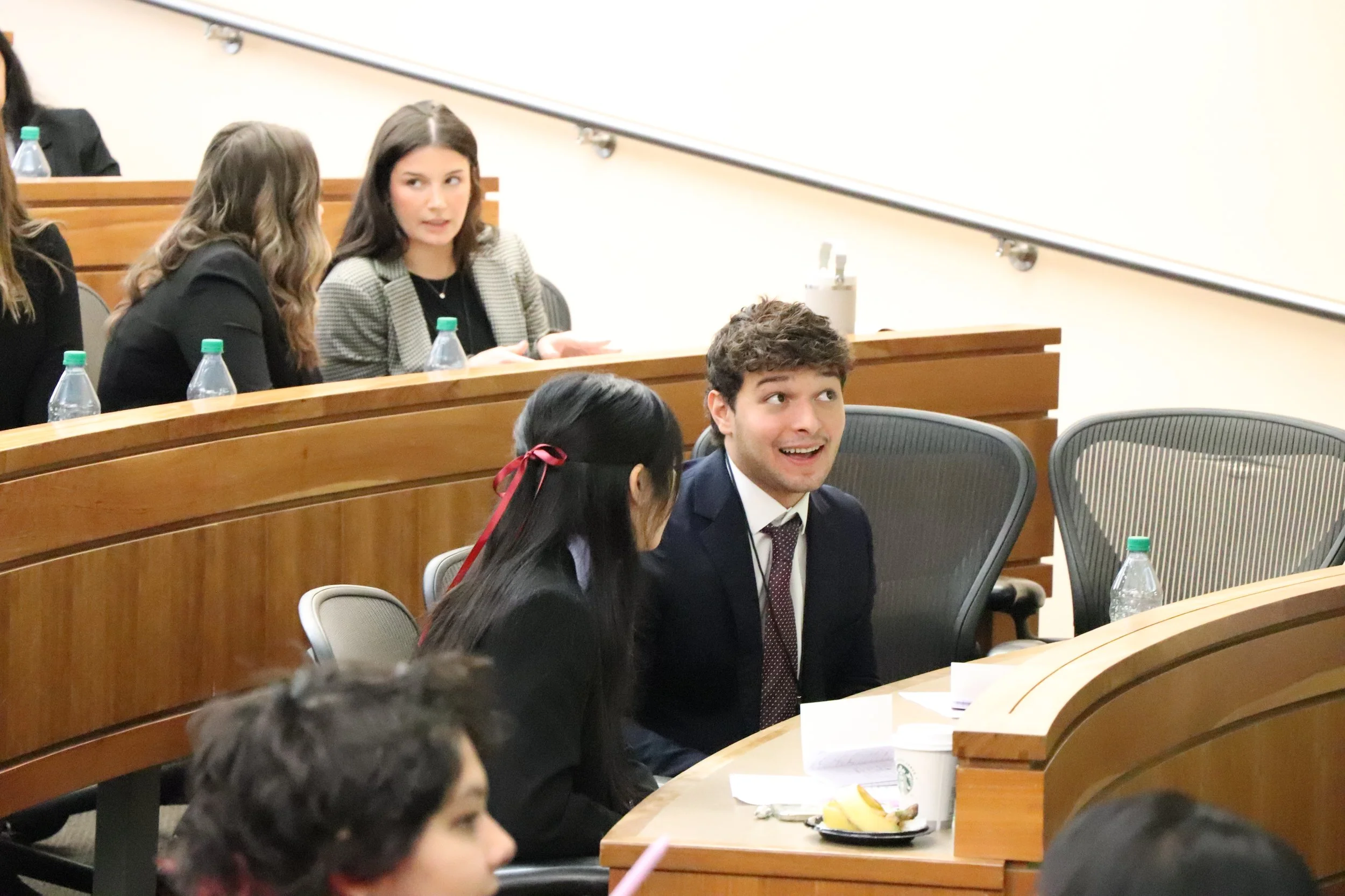 People seated in a classroom or conference room, engaging in a discussion, with water bottles, papers, and food on the tables.