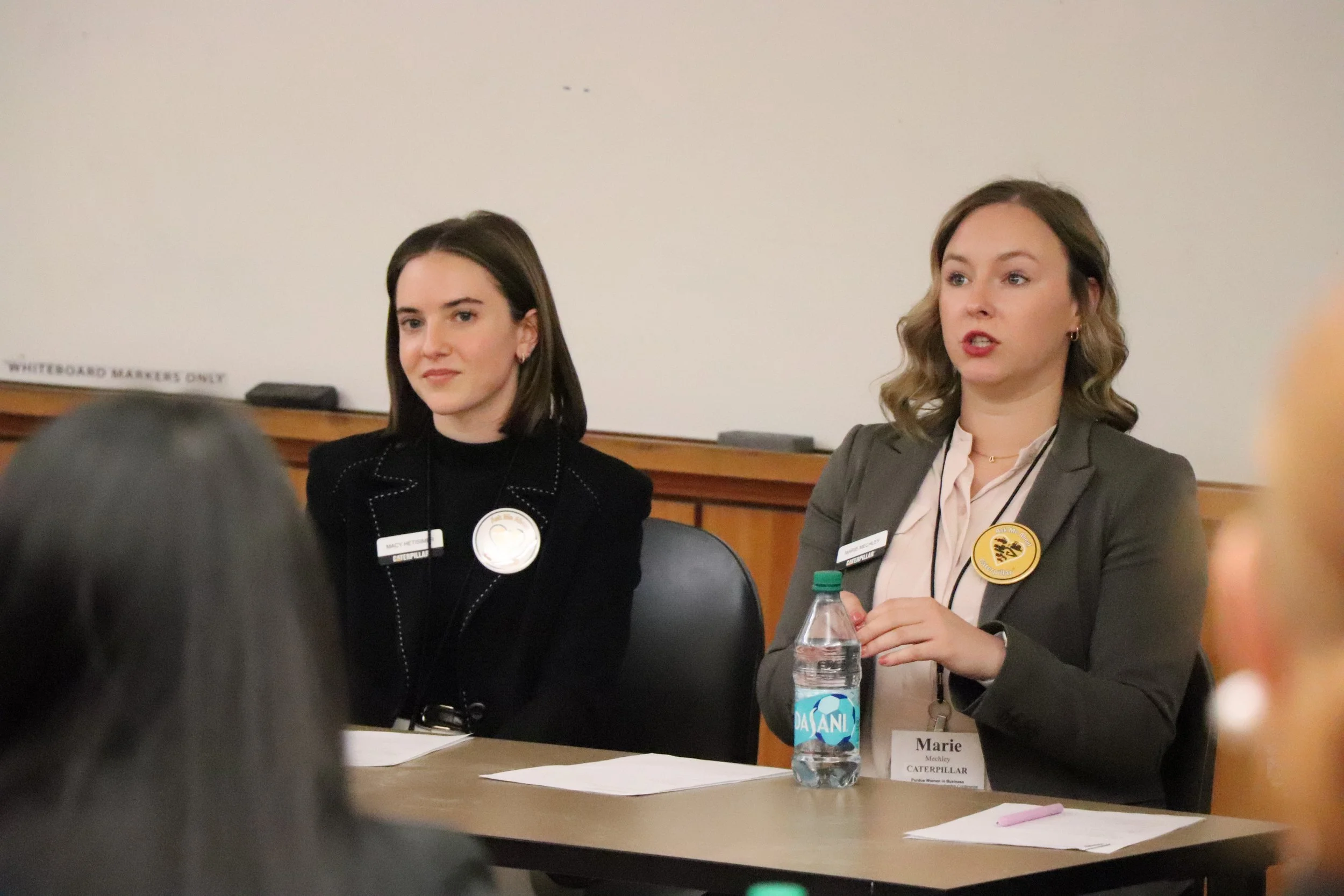Two women sitting at a panel table during an event, one with dark hair in a black blazer and the other with light brown hair in a gray blazer, both wearing name tags, with papers and a water bottle on the table.