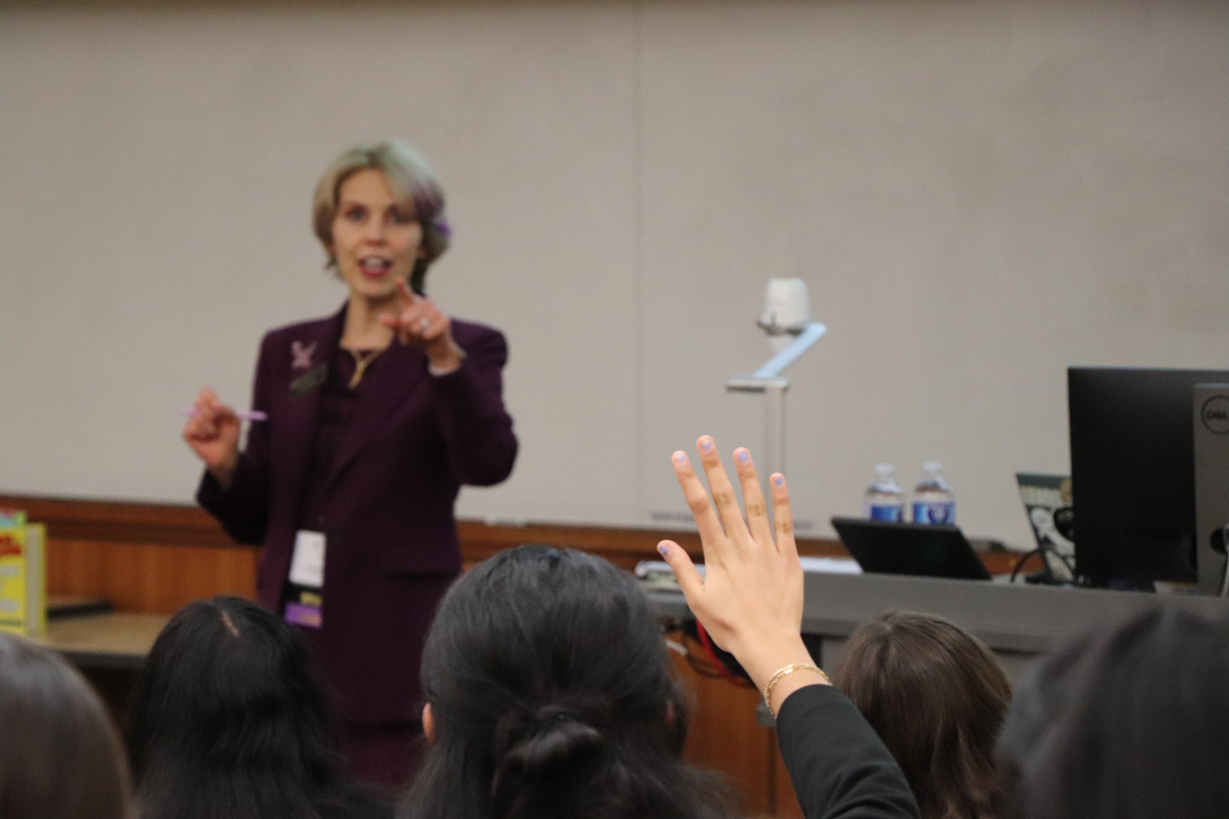 A woman in a dark purple blazer speaking to an audience, with one audience member raising their hand, in a conference room setting.
