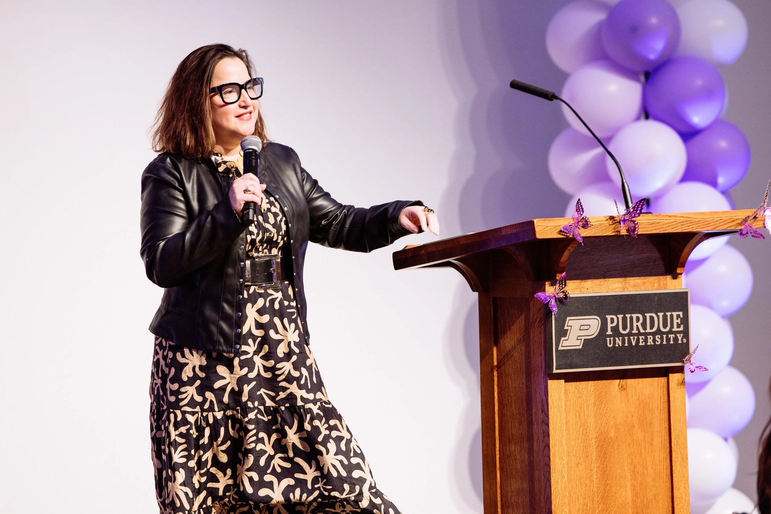 Woman wearing glasses and a black leather jacket giving a speech at a podium with Purdue University logo, decorated with purple butterfly cutouts and a backdrop of purple and white balloons.