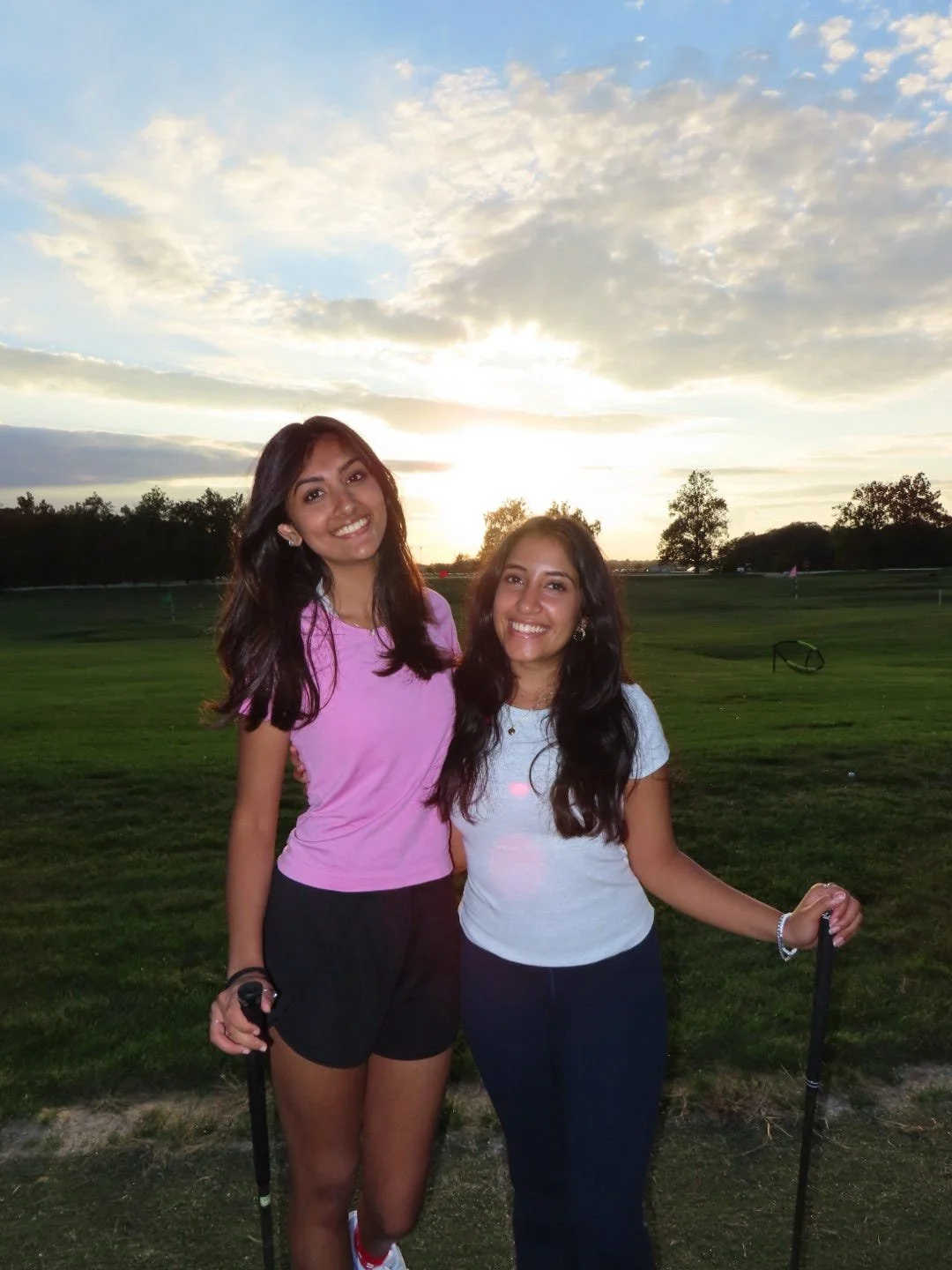 Two young women standing together on a golf course at sunset, smiling, with trees and a partly cloudy sky in the background.