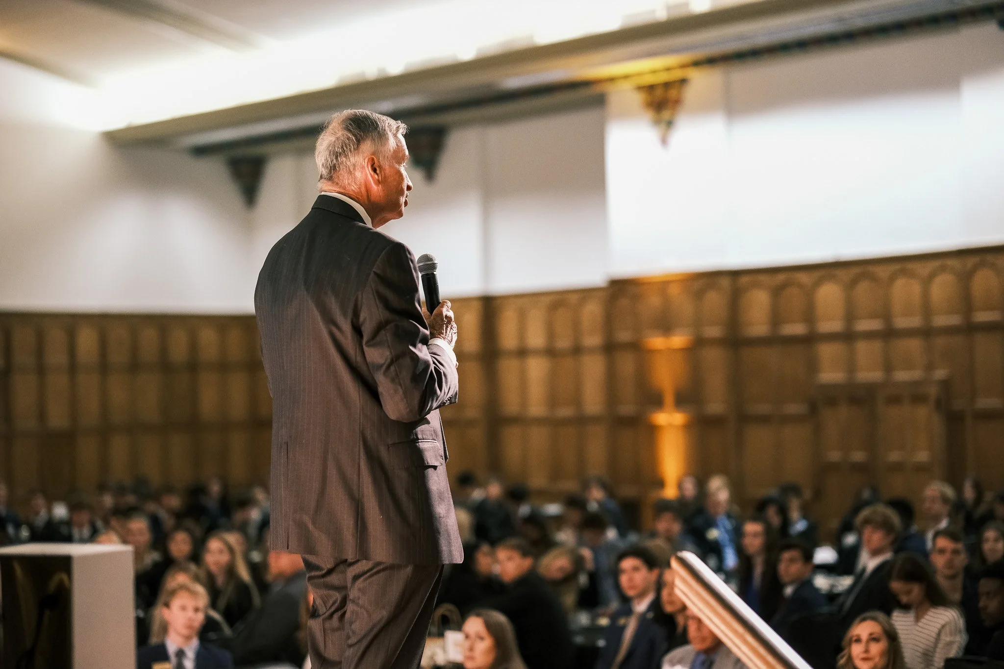 Older man in a suit holding a microphone speaking to an audience in a large room with wooden walls and a high ceiling.