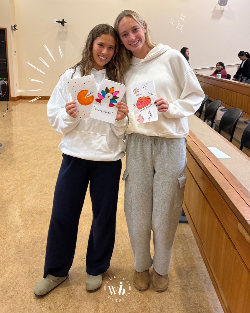 Two young women standing in a classroom, smiling, each holding a piece of art. The woman on the left has curly hair and is holding a drawing of a crescent moon, while the woman on the right has straight hair and is holding a drawing of a red clutch with flowers around it. In the background, there are other people seated and walking, with a table to the right and a wall with some decorations.