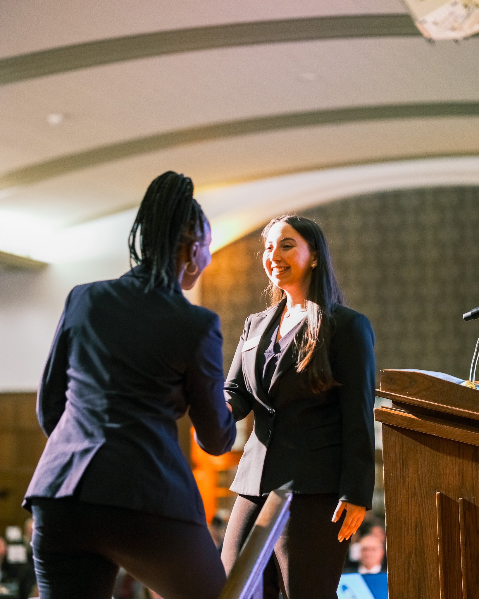 Two women in business attire shaking hands on stage during an award or recognition ceremony in a conference hall.