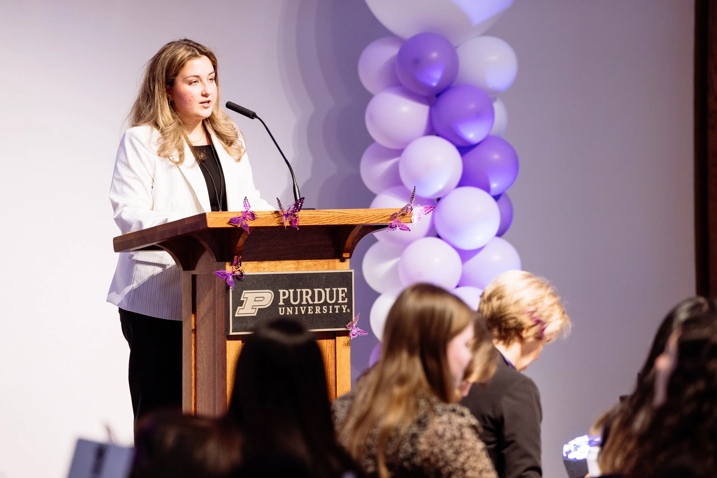 A woman in a white blazer and black shirt is speaking at a podium with a Purdue University sign, decorated with purple butterflies, in front of a balloon arrangement of white and purple balloons.