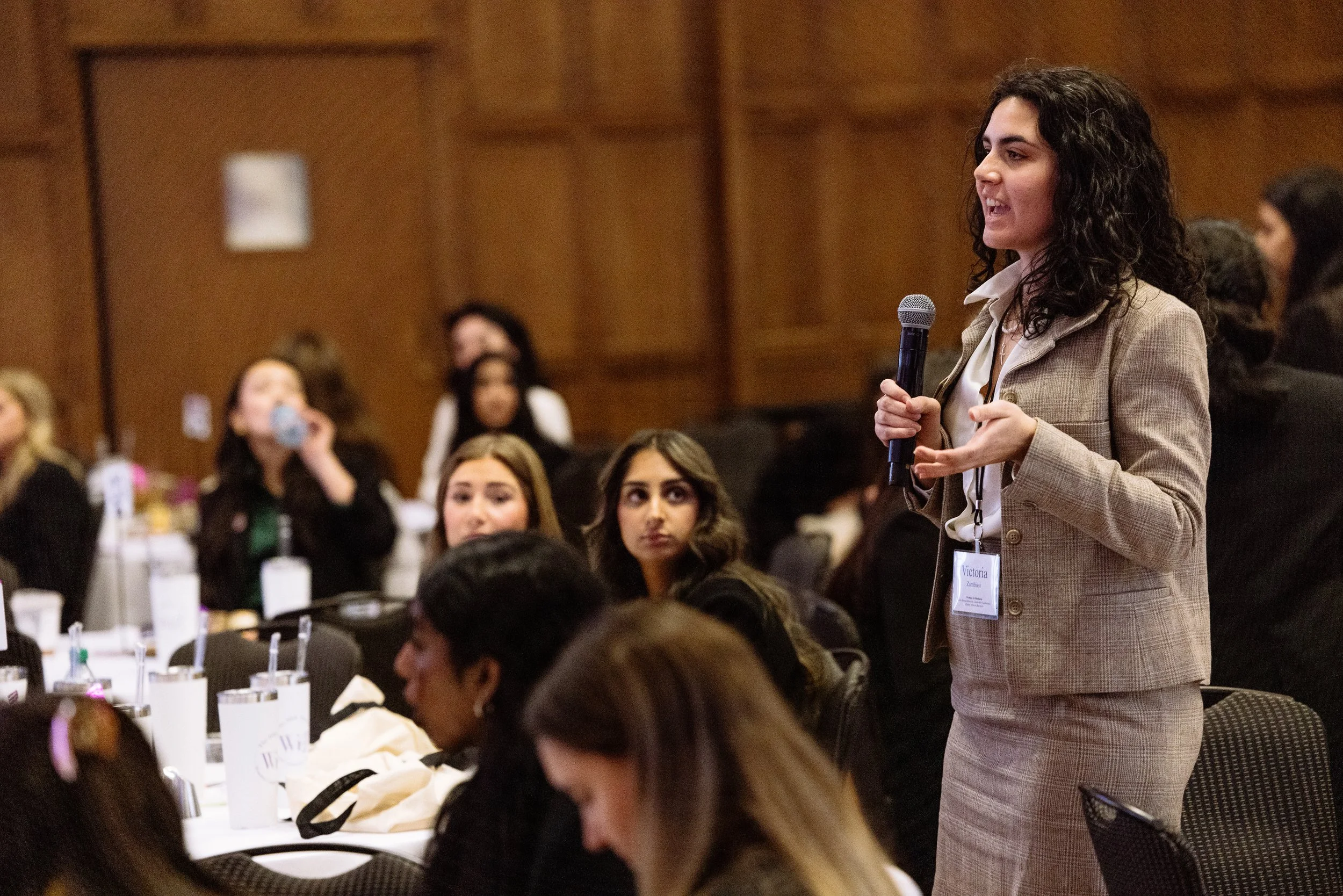 A woman with curly dark hair wearing a beige blazer and skirt speaking into a microphone at a conference. Several women are seated at tables and listening, some looking at her and others looking away or drinking water. The room has wood-paneled walls