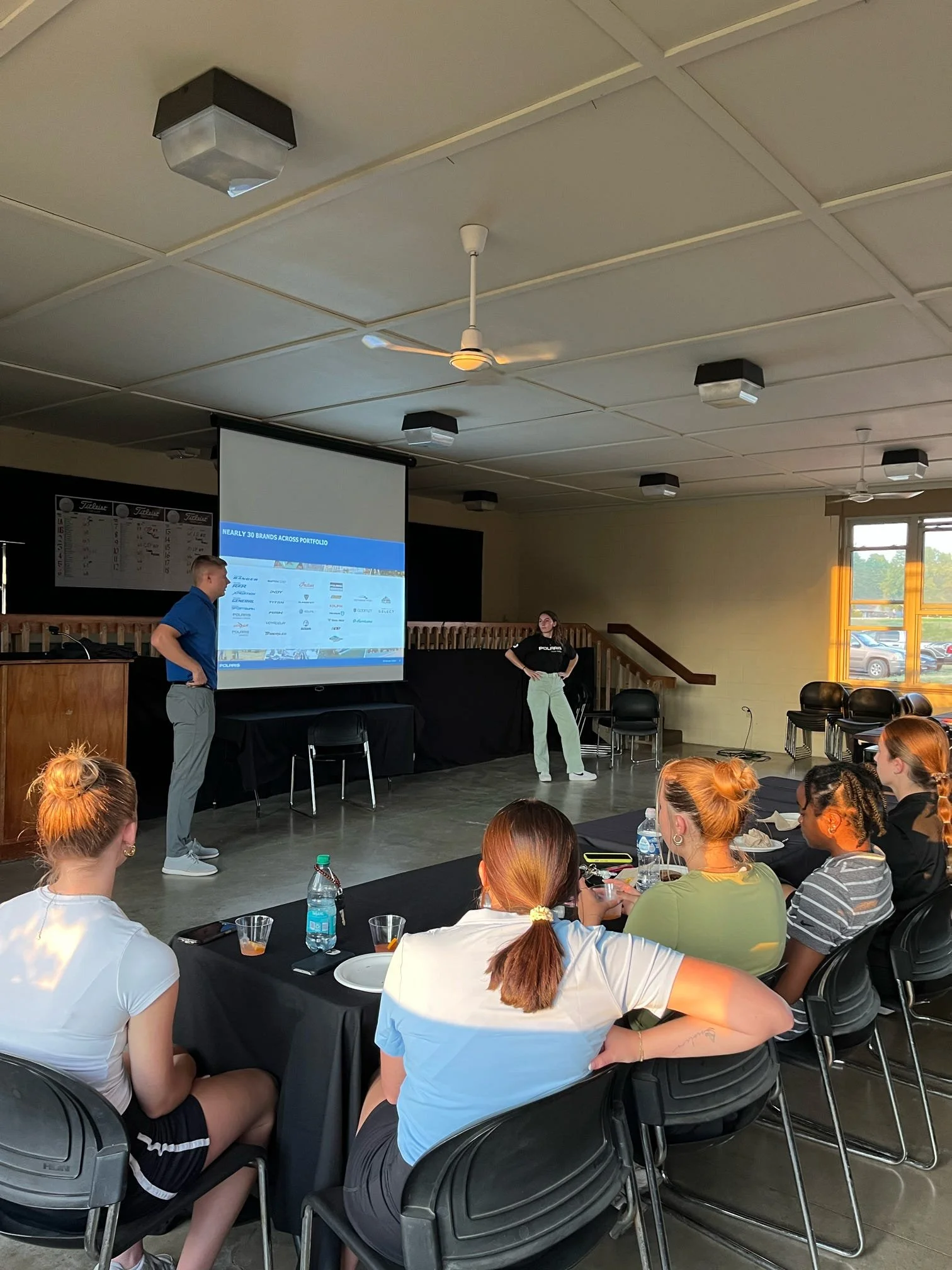 A person is giving a presentation on a screen in a room with several seated women, some with drinks and bottled water on the table, during daytime with natural light coming through a window.