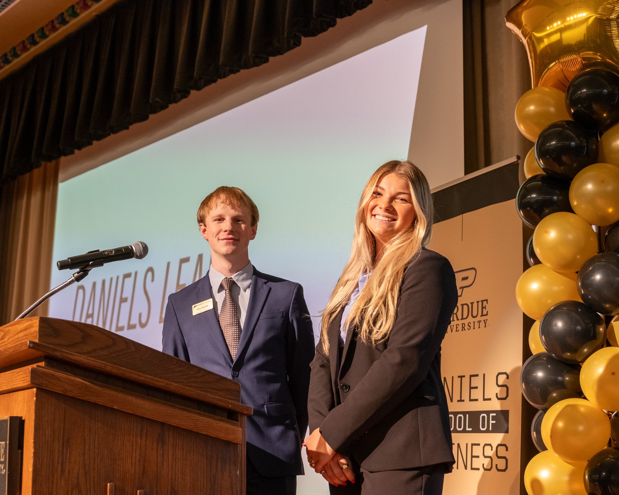 A young man and a woman standing on stage at an event, smiling. The young man is wearing a navy suit and tie, and the woman is in a black blazer. There is a podium with a microphone in front of them, and a large screen behind displaying the name 'DANIELS' and 'SCHOOL OF BUSINESS'. To the right, there are gold and black balloons. An event banner indicates it is at Purdue University.