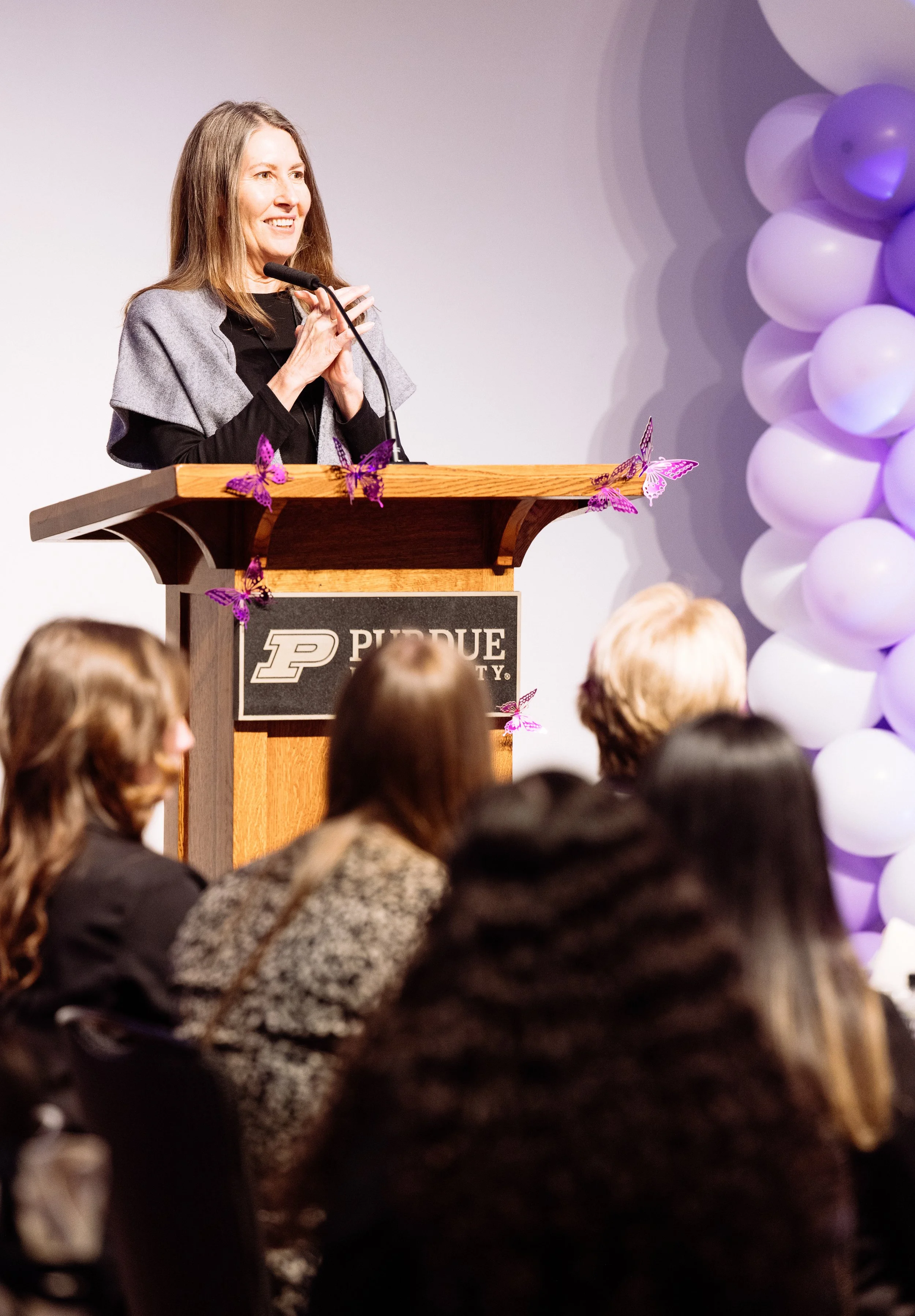A woman speaking at a project or event at Purdue University, standing behind a wooden podium with purple butterfly decorations, while an audience listens.