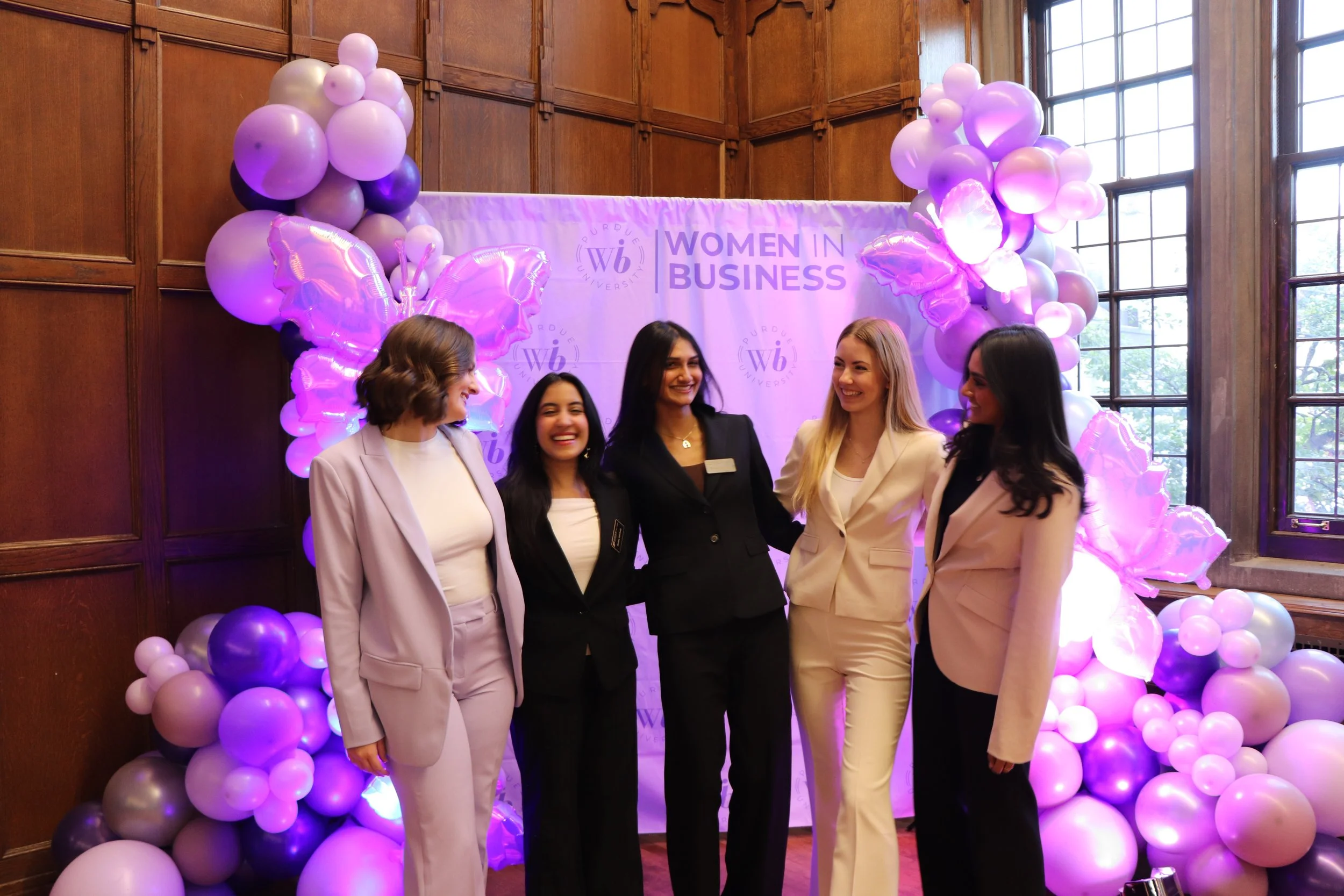 Five women in business attire smiling and posing together at a Women in Business event with purple balloon decorations and a purple backdrop.
