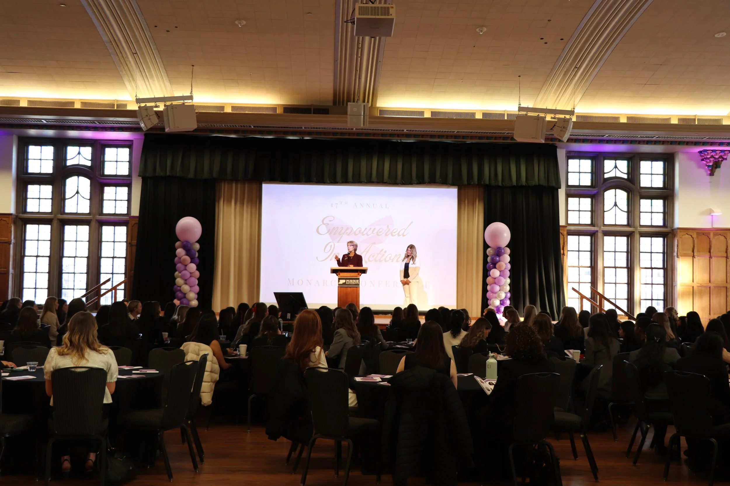 Conference in a large room with attendees sitting at tables, a stage with two women speaking, and balloon columns on either side of the stage.