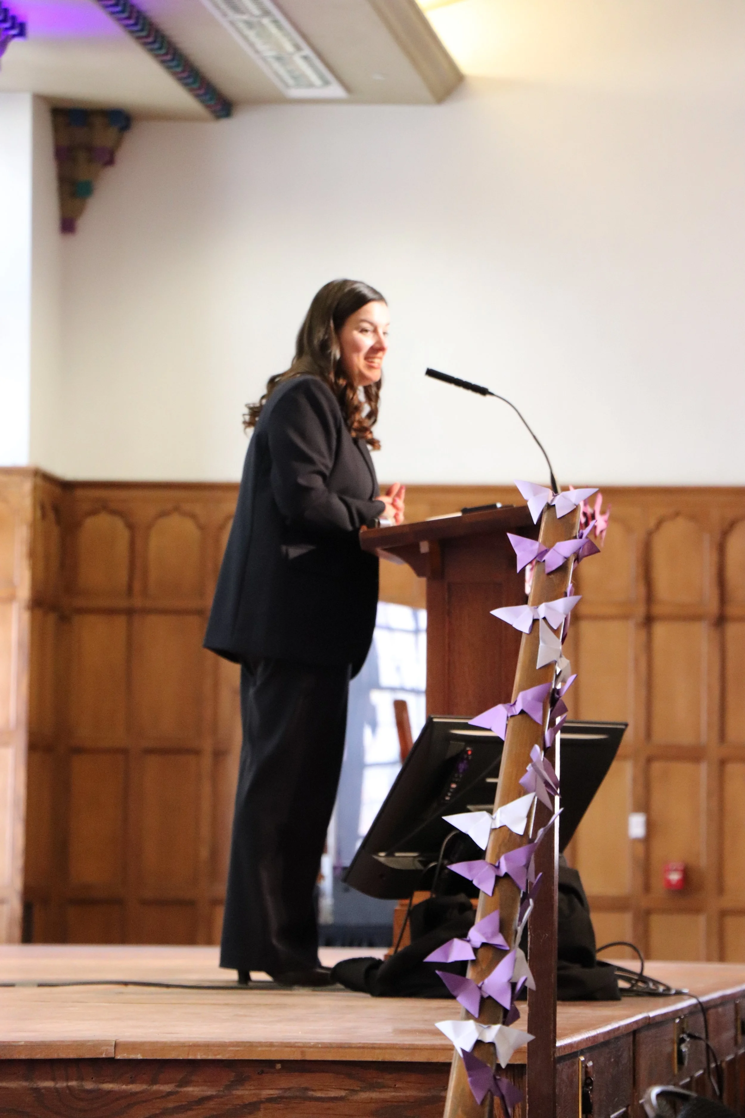 A woman in a black suit standing at a wooden podium decorated with purple and white paper butterflies, speaking into a microphone in a wood-paneled room.