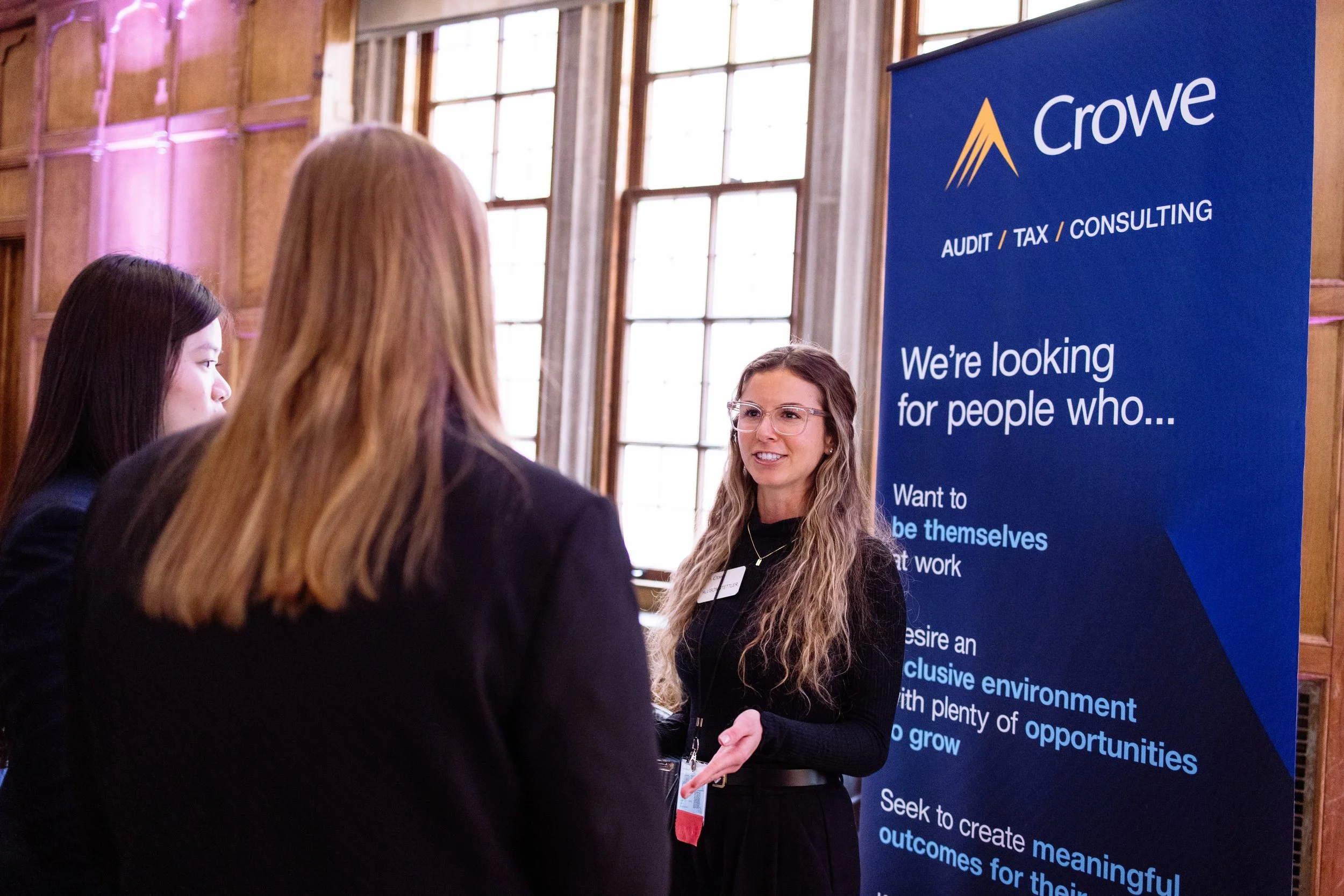 Three women in conversation at a professional event, with a blue banner behind them displaying the logo 'Crowe' and text about auditing, tax, and consulting services.