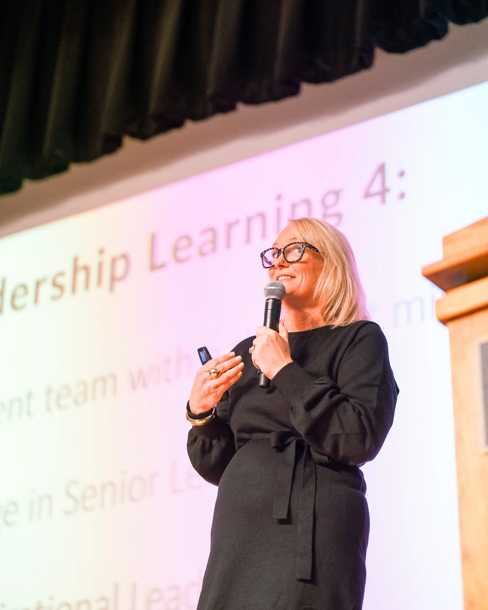 A woman with blonde hair and glasses giving a presentation on stage, holding a microphone and a remote clicker, with a slide that reads 'Leadership Learning 4:' in the background.