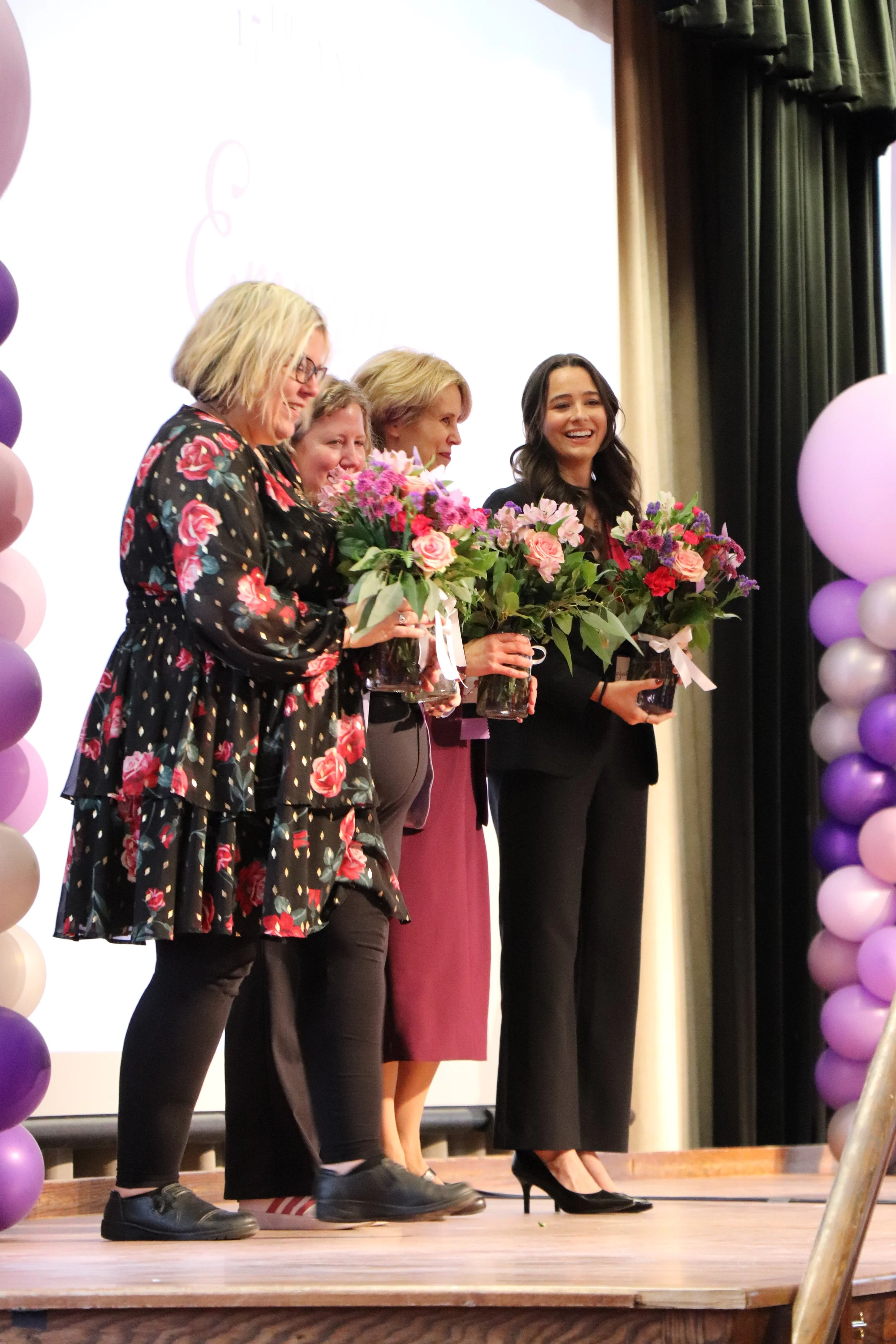 Four women standing on a stage holding colorful flower arrangements, with purple and pink balloon decorations on either side.
