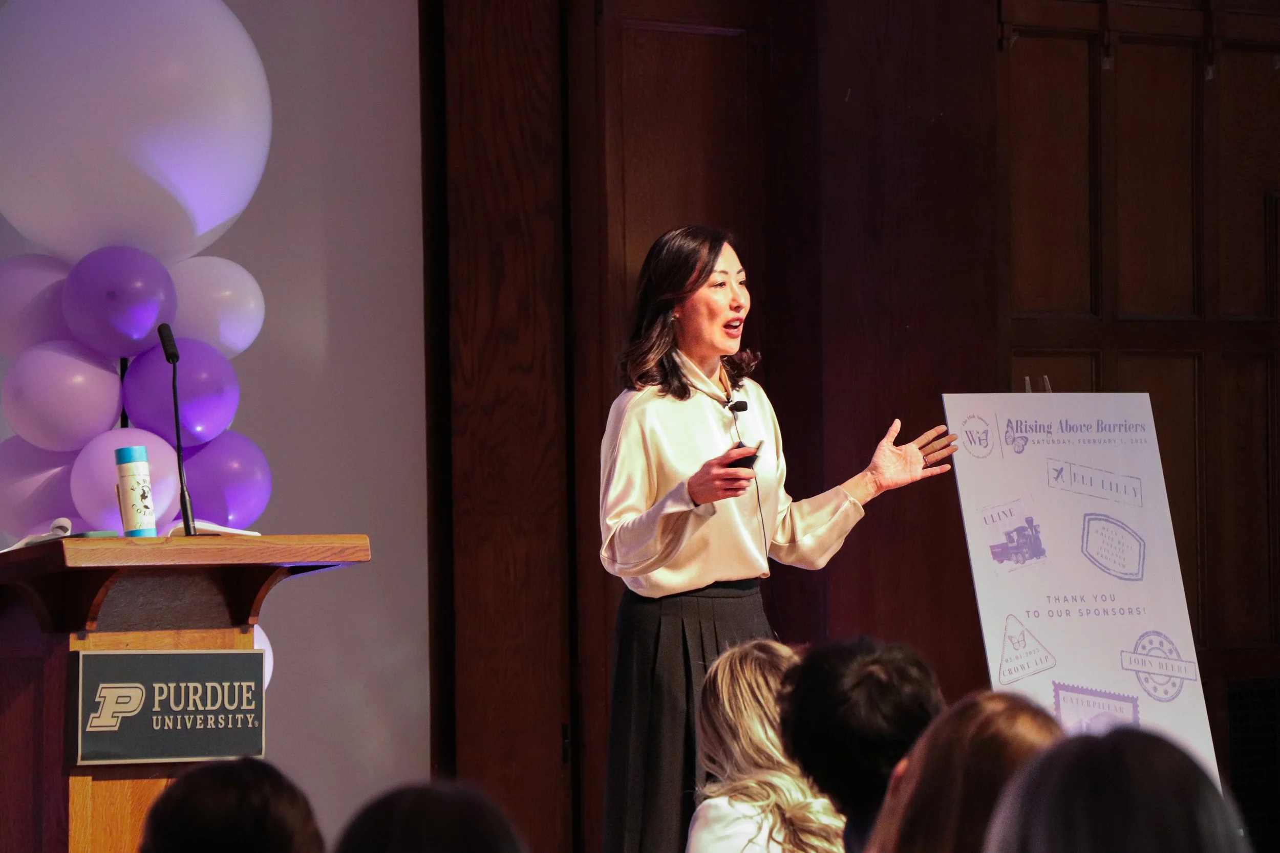 Woman giving a presentation at Purdue University, standing near a poster with illustrations and text, with a podium adorned with purple balloons and a hand sanitizer bottle.