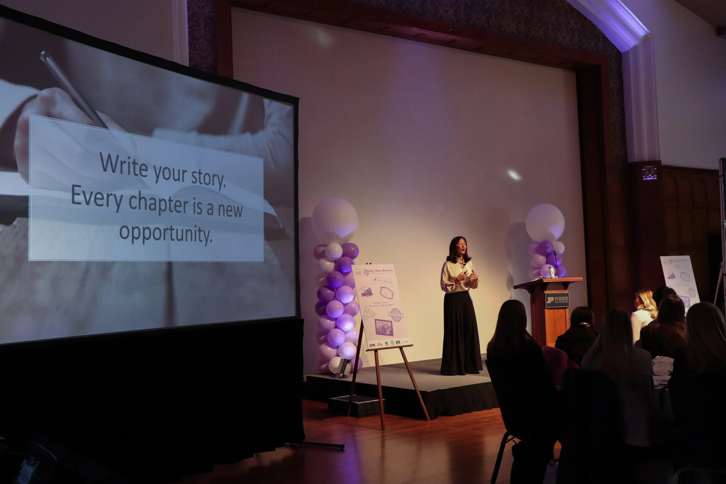 A woman standing on stage at Purdue University giving a presentation, with a large screen displaying a message that says, 'Write your story. Every chapter is a new opportunity.' The stage is decorated with purple and white balloons.