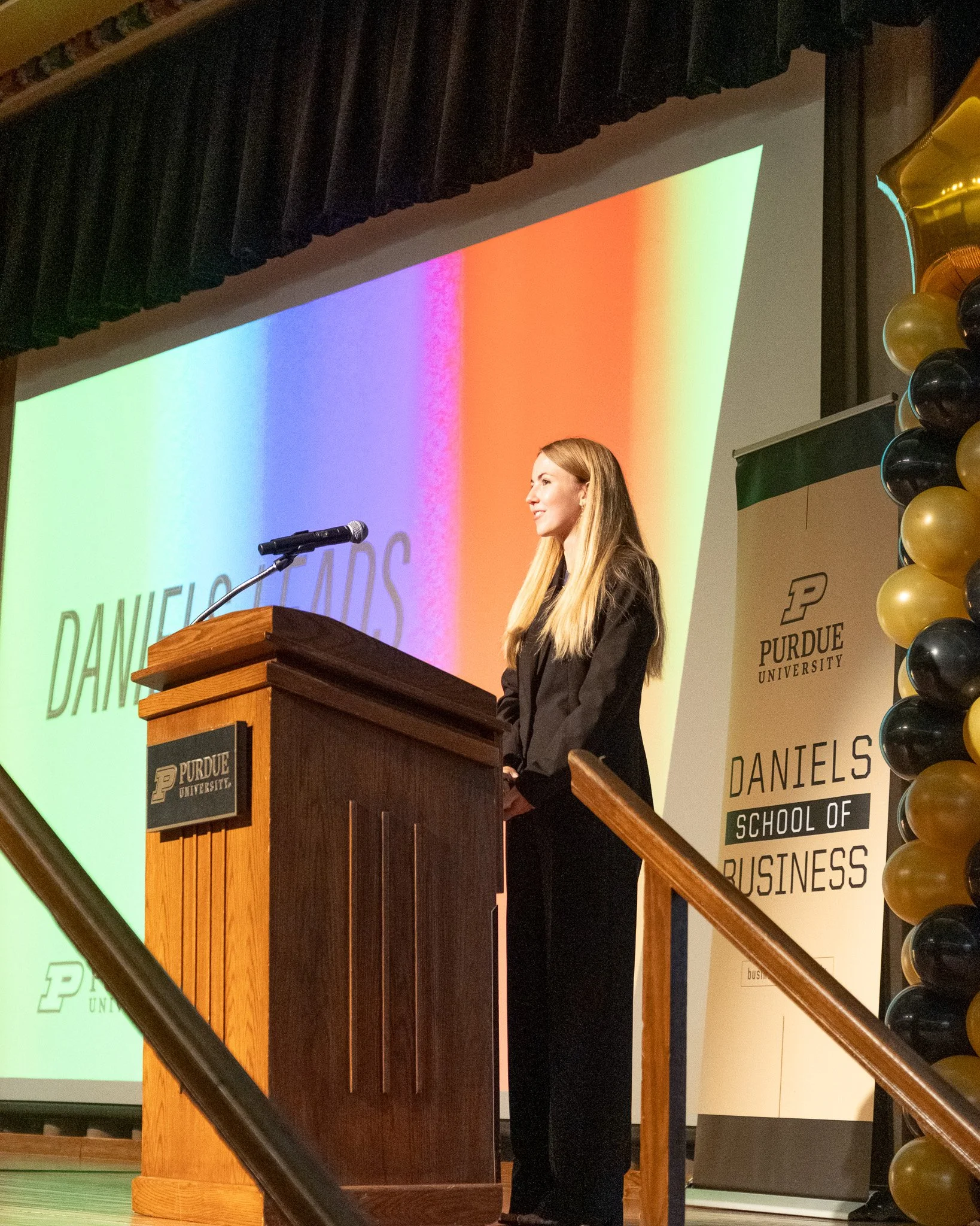 Young woman standing at a podium with a Purdue University logo, delivering a speech or presentation at Daniels School of Business event, with a colorful backdrop and balloon decorations.