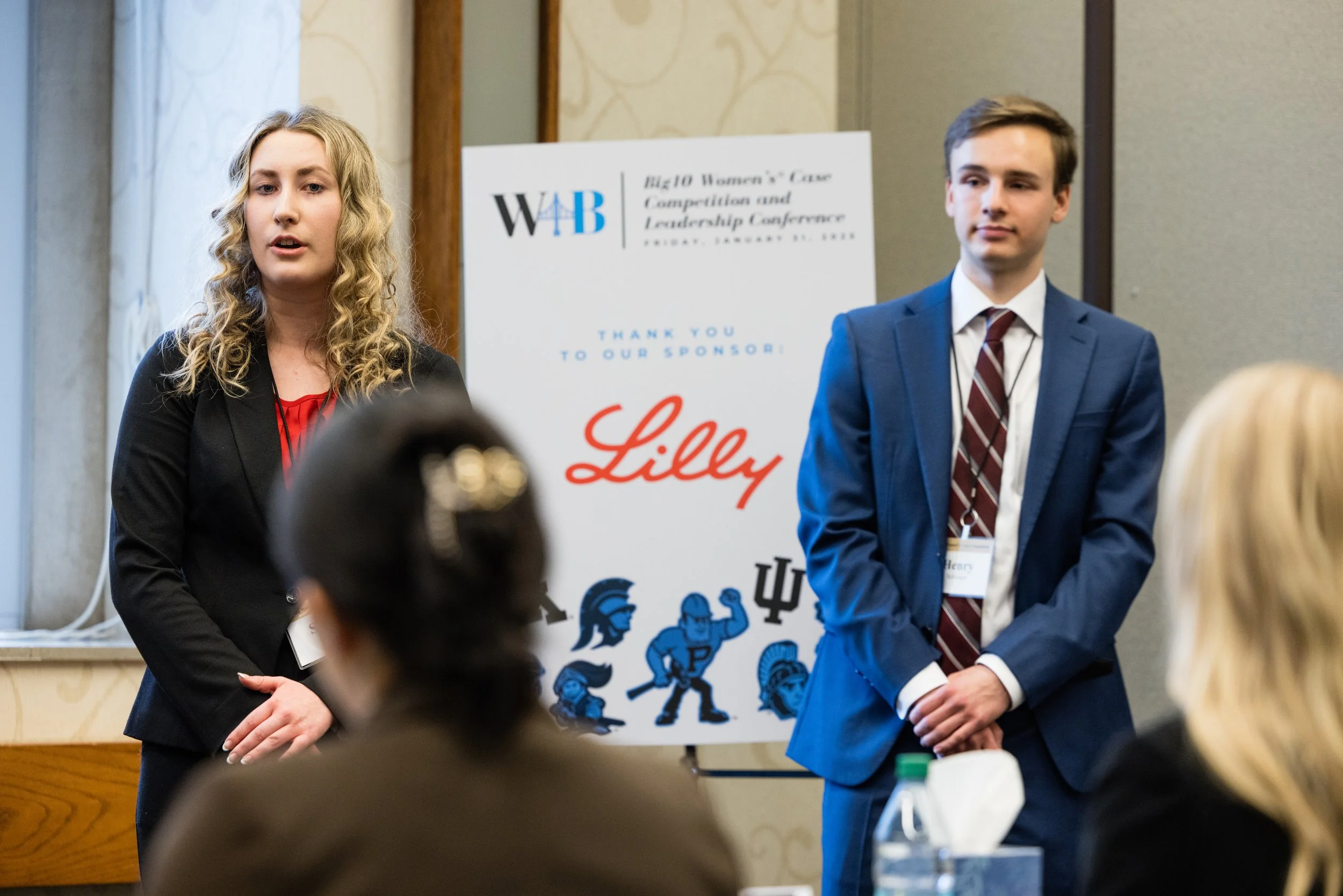 Two young professionals, a woman with curly blonde hair and a man with short brown hair, stand in front of a poster at a conference. The woman is speaking and the man is listening. The poster behind them reads 'Big Ten Women's Care Competition and Le