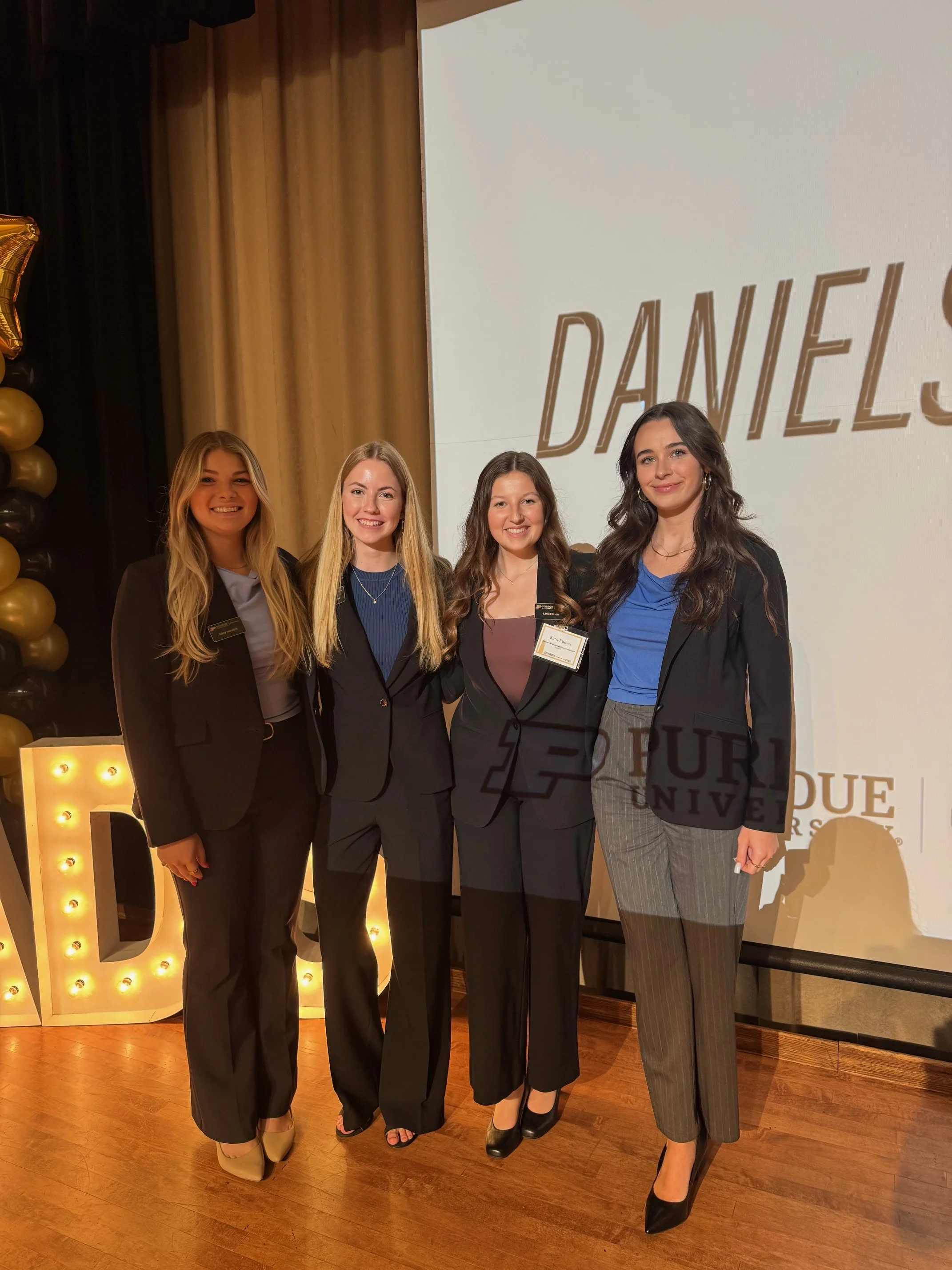 Four women dressed in formal business attire standing together on a stage, smiling, with a large projection screen behind them displaying the name 'DANIELS' and 'Purdue University' logo. The stage is decorated with gold and black balloons and a large illuminated letter 'D'.