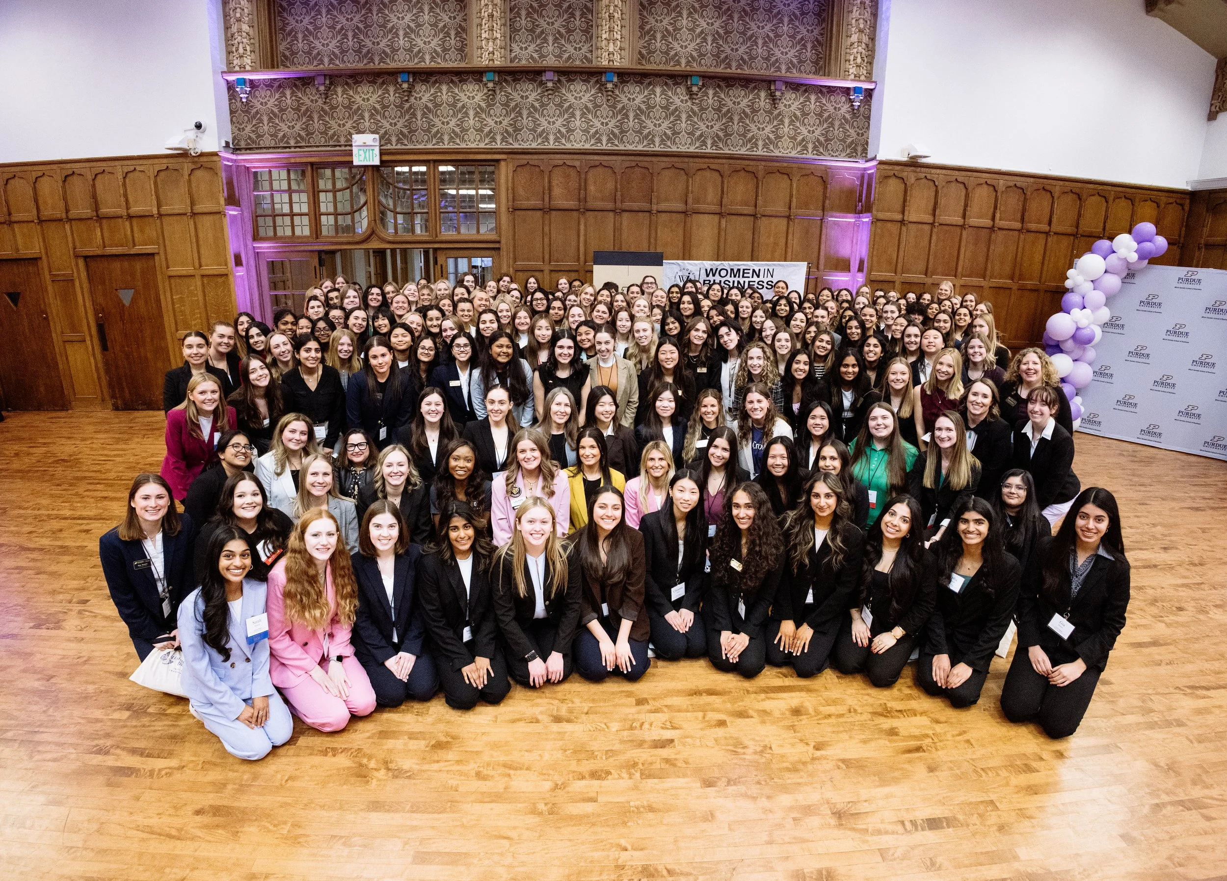 Large group of women in professional attire gathered in a ballroom with wood-paneled walls and a wooden floor, attending a 'Women in Business' event. They are smiling at the camera, with balloons and a banner in the background.