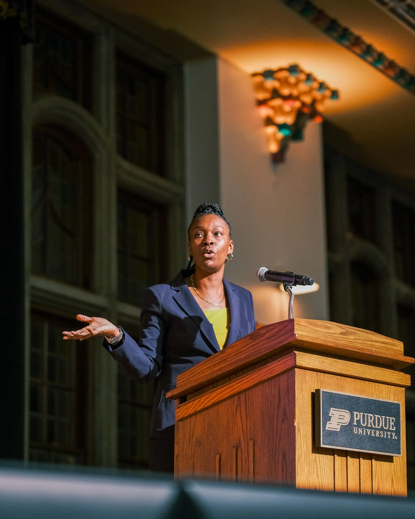 A woman in a navy blazer and yellow top speaking at a podium with a Purdue University sign, gesturing with her hands, in a large room with stained glass windows.