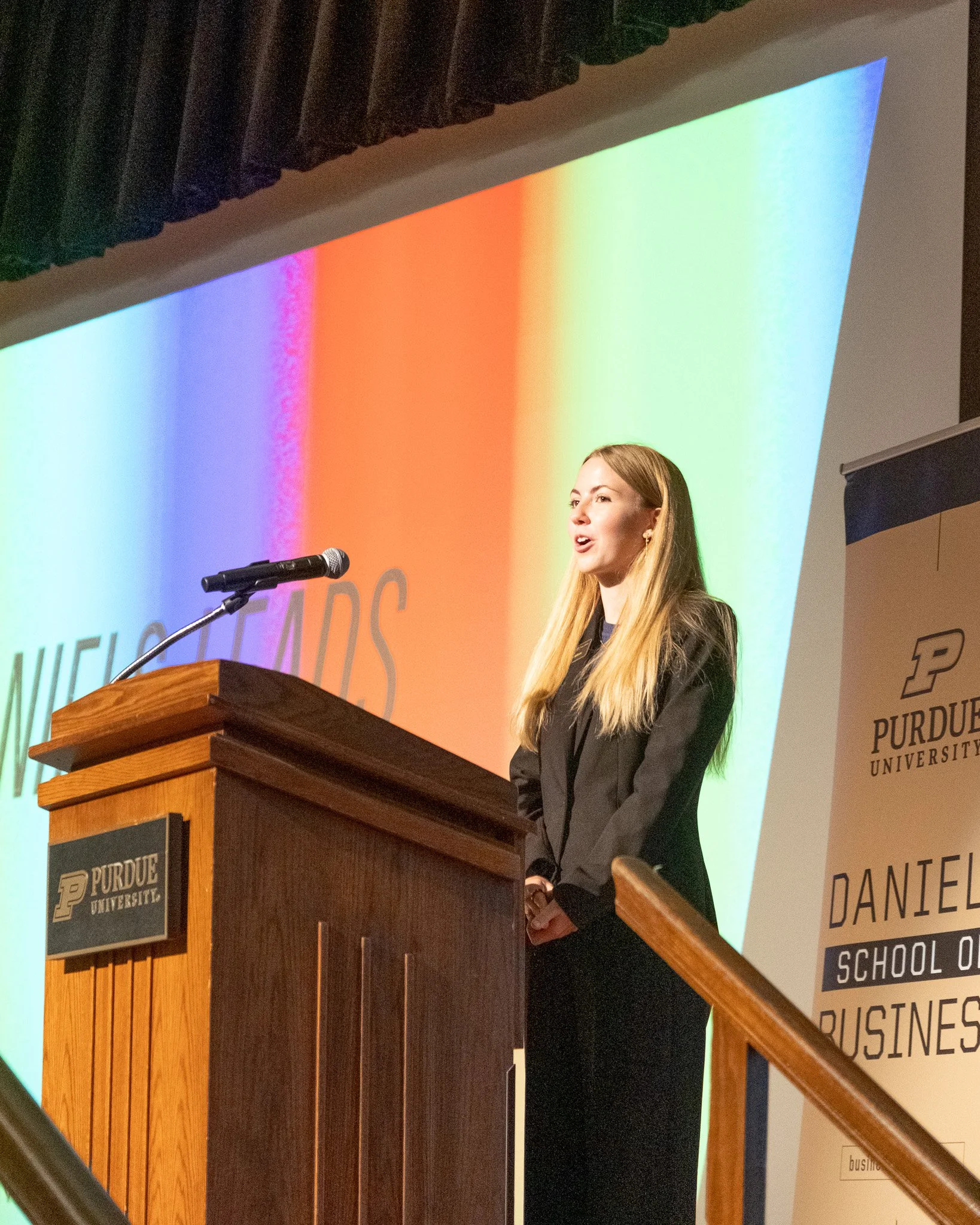 A young woman with long blonde hair is standing at a wooden podium with a Purdue University logo. She is speaking into a microphone in front of a colorful rainbow background on a large screen. A banner with the Purdue University logo and text is visible on the right side.