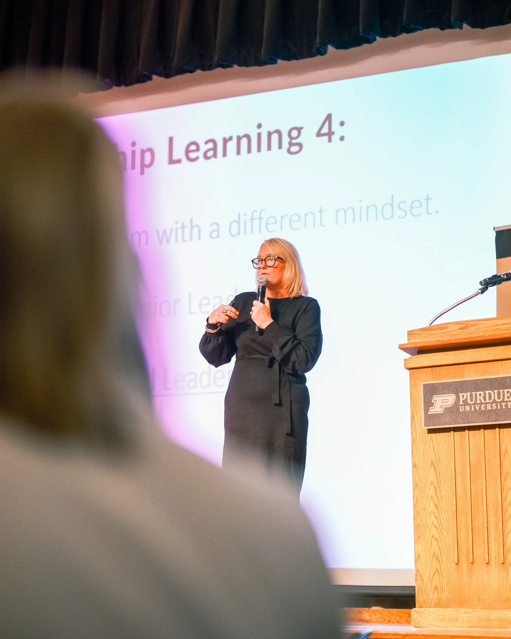 A woman in black standing on stage at Purdue University, holding a microphone, giving a presentation with a slide that reads 'Leadership Learning 4:'.
