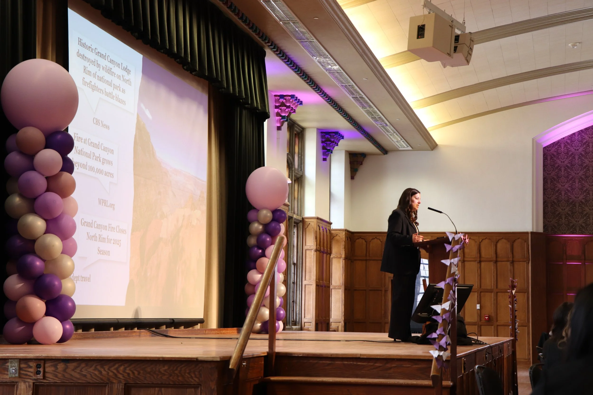 A woman speaking at a podium on a stage decorated with balloon columns in shades of purple, pink, and gold, in a wood-paneled room with a large projection screen displaying information about Grand Canyon National Park fire closures.