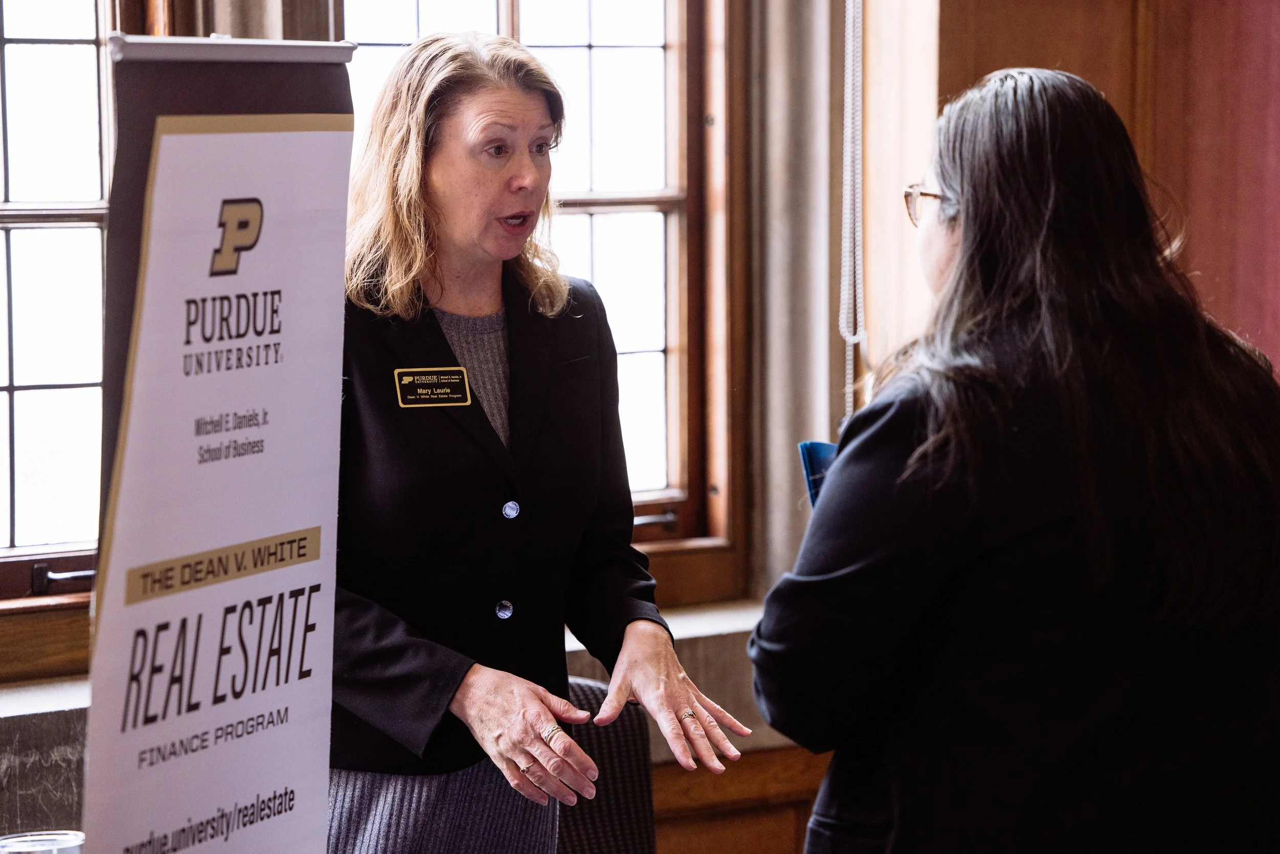 A woman with a name tag speaking with a woman with long dark hair at a Purdue University event, near a sign for the Dean V. White Real Estate Finance Program.