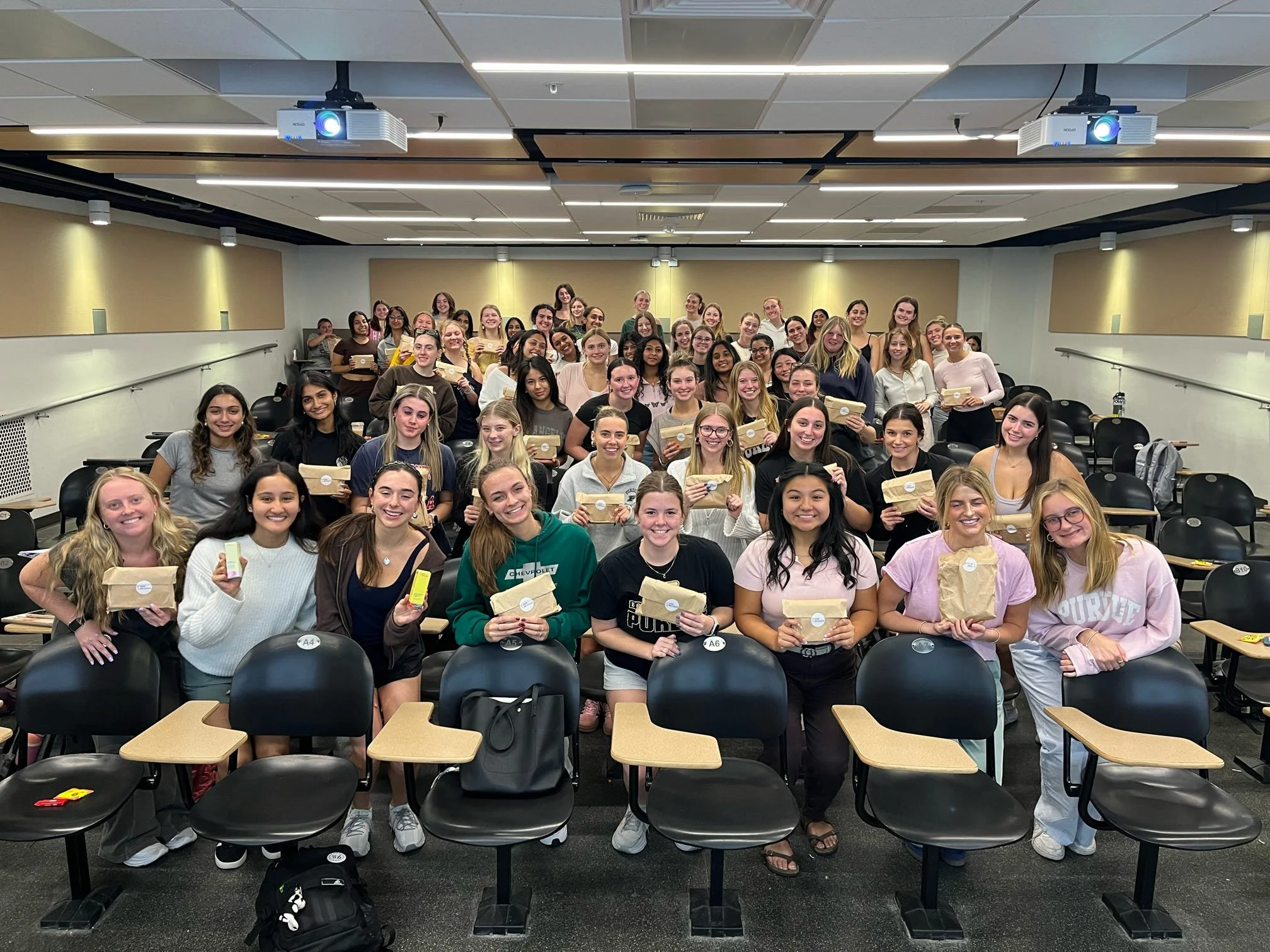 A large group of young women in a classroom or lecture hall holding what appear to be awards or gifts, smiling at the camera.