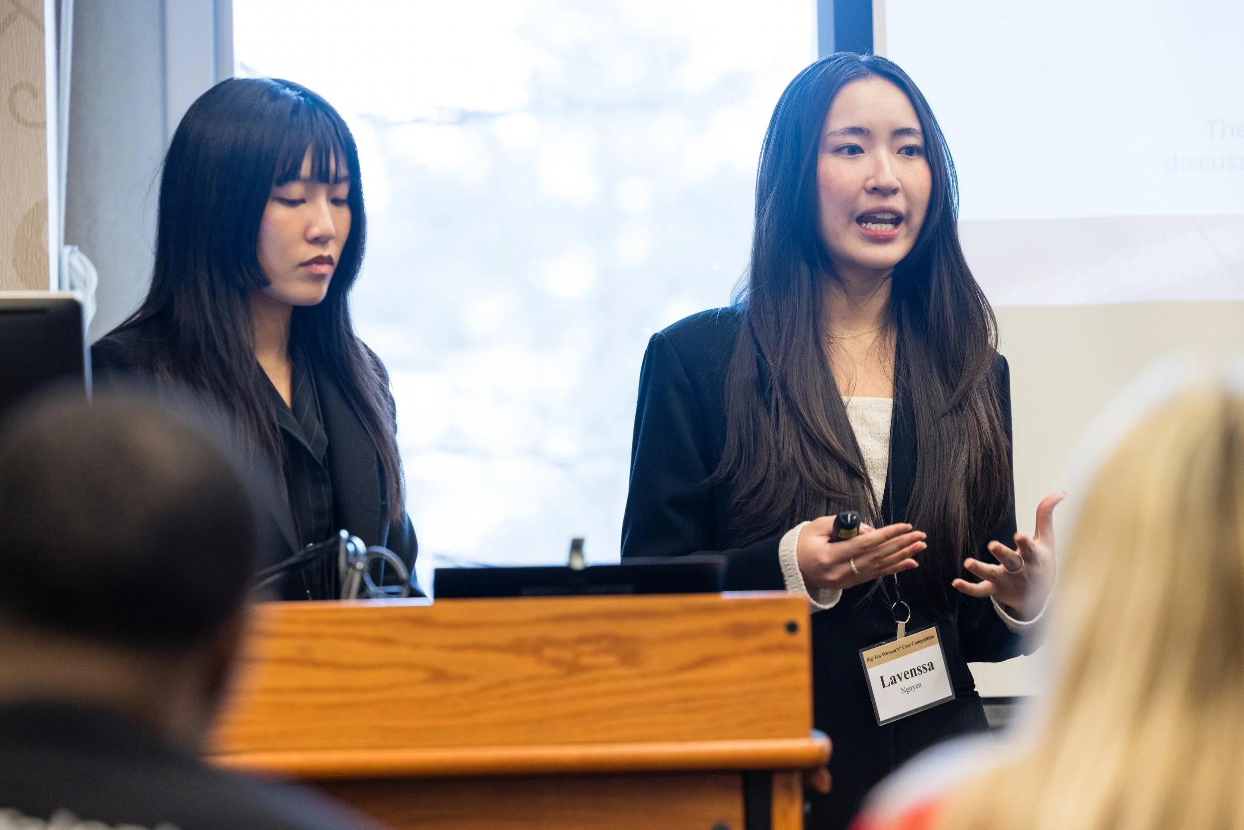Two women presenting in a conference room, one speaking and the other looking down, with audience members in the foreground.