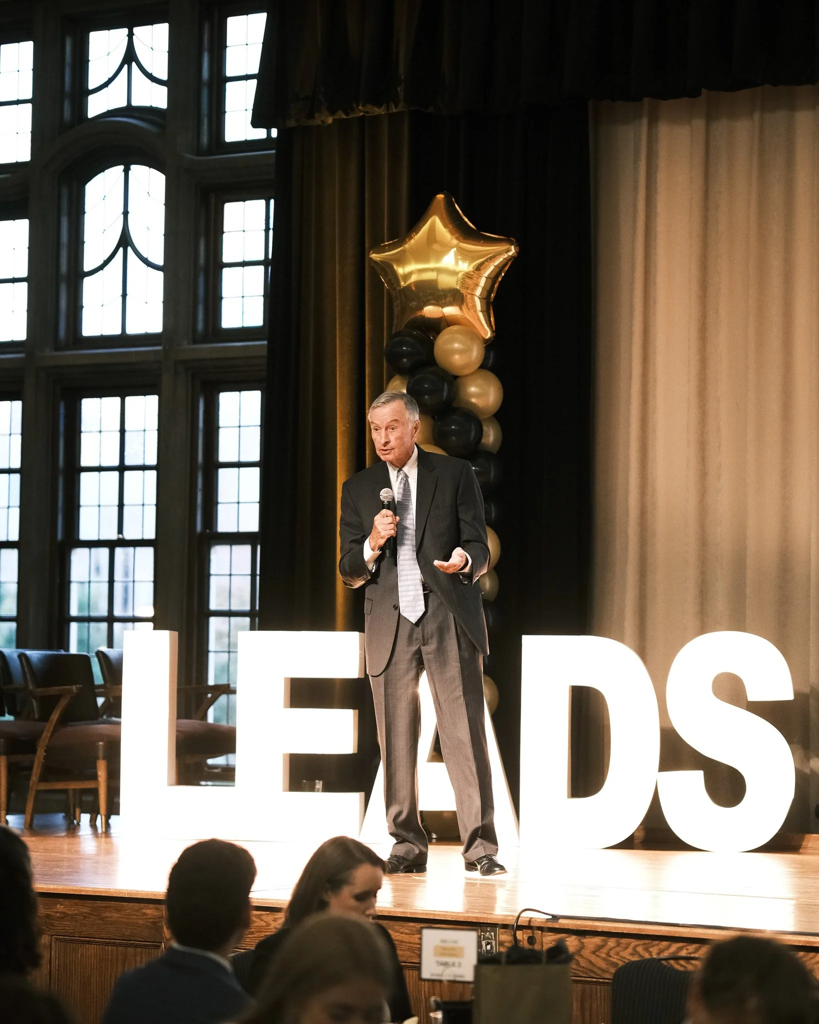 A man in a suit speaking into a microphone on stage with large illuminated letters spelling 'LEADS' behind him, in a venue with tall windows and black and gold balloons.