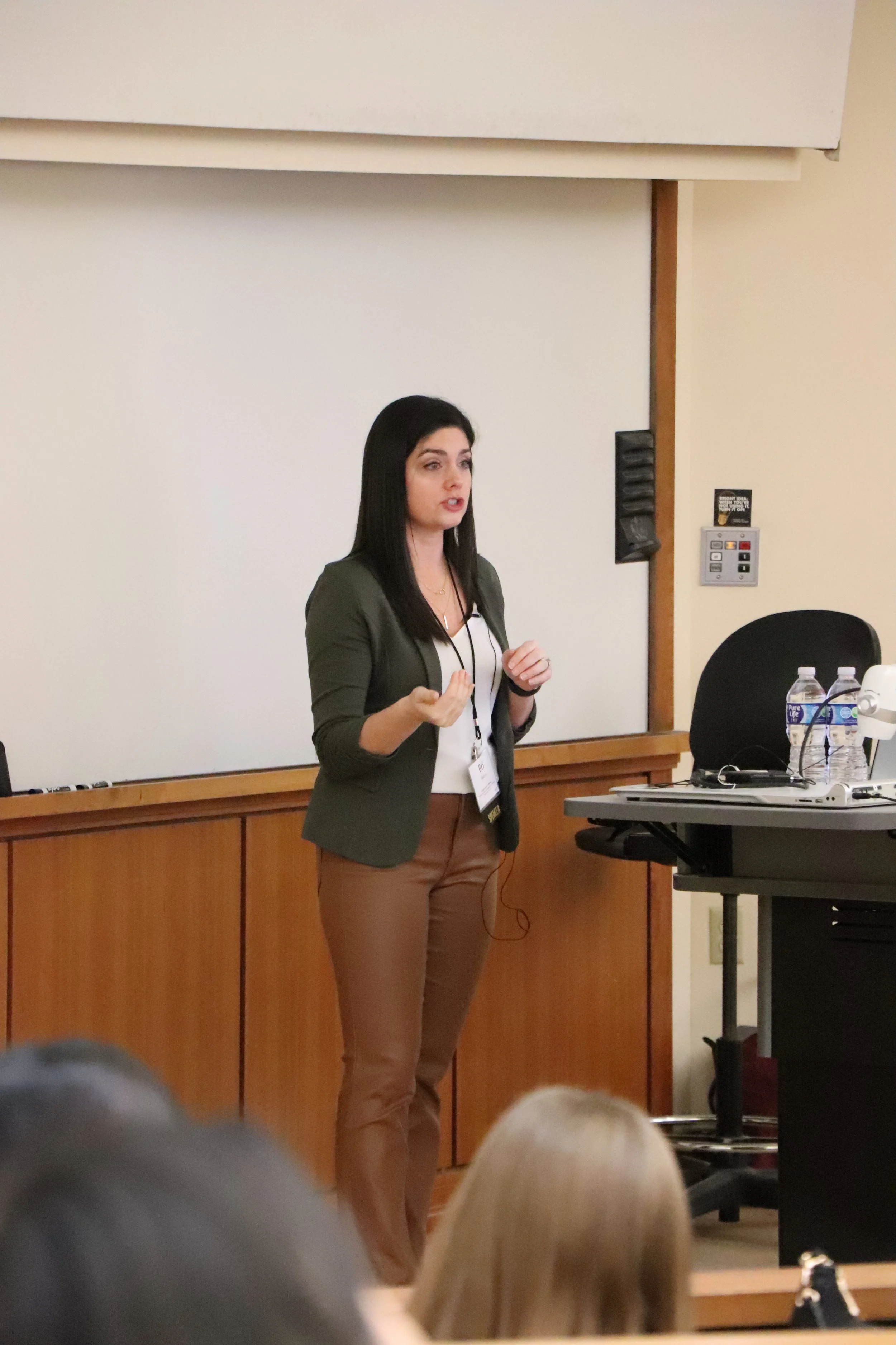 A woman giving a presentation in a classroom or conference room, standing in front of a whiteboard with a projection screen, with audience members visible in the foreground.