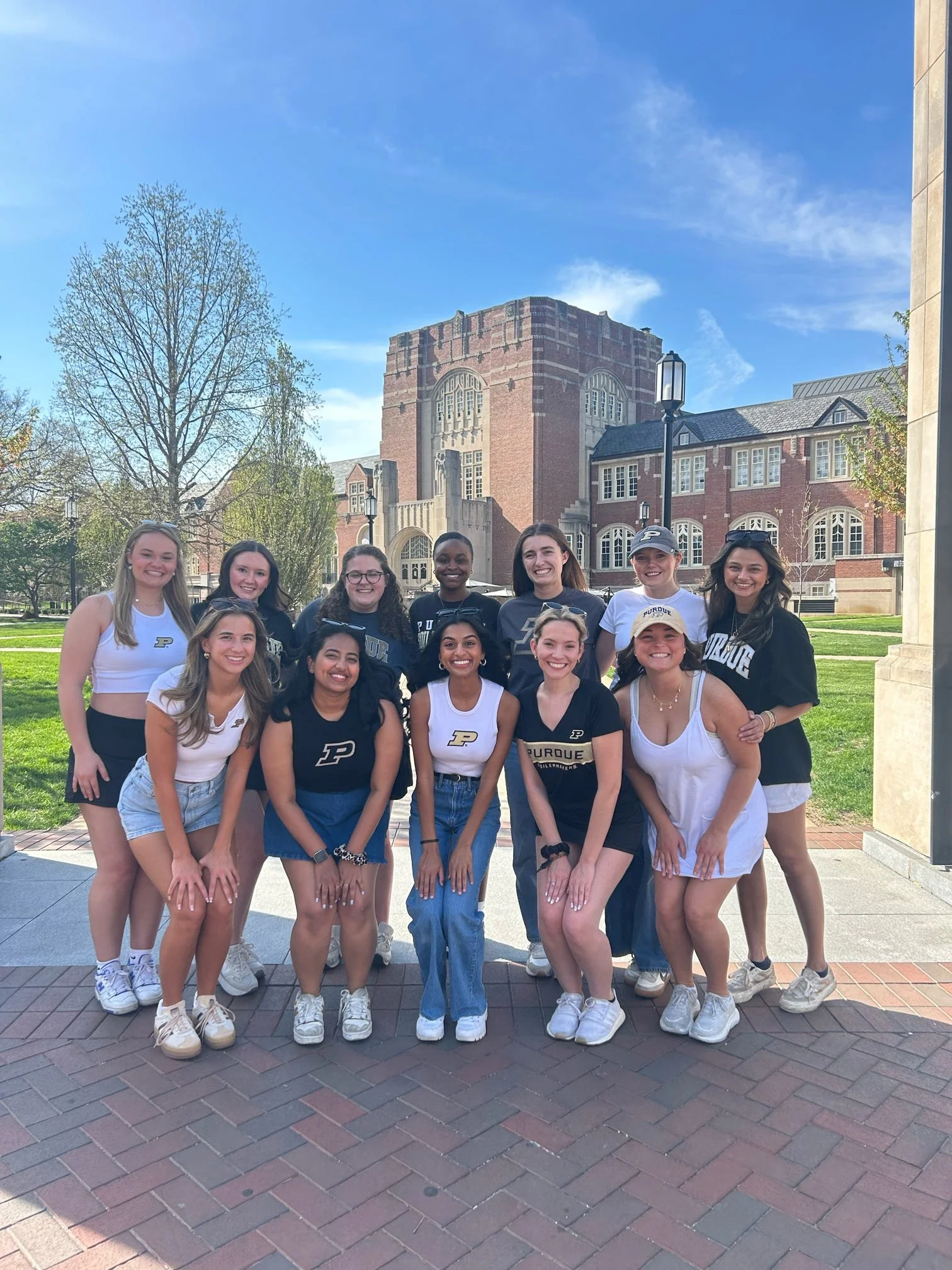 Group of young women posing outdoors on a sunny day, wearing Purdue University apparel, with a historic campus building in the background.