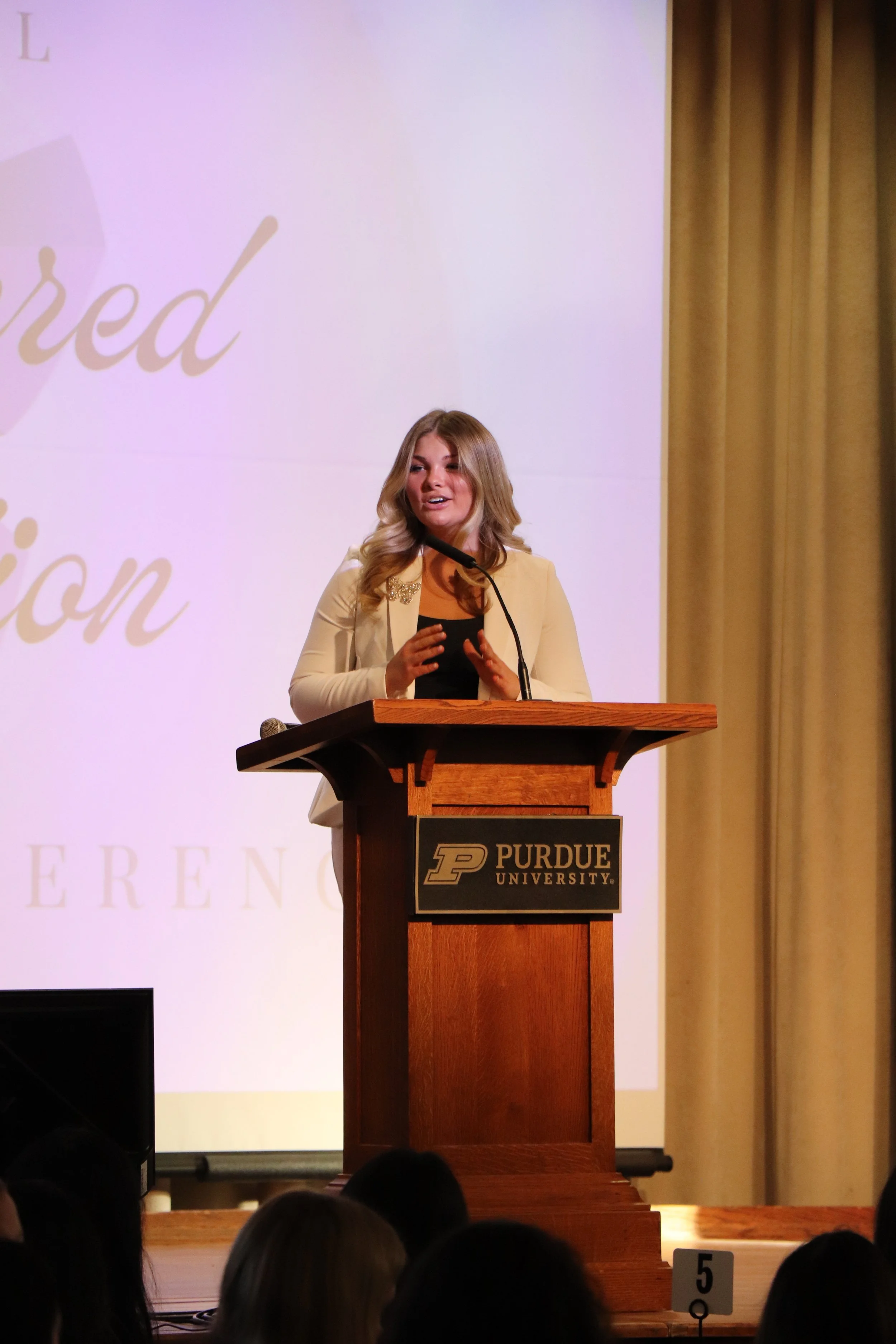 A young woman with blonde hair speaking at a podium with a microphone at Purdue University. She is wearing a white blazer and a black top. Behind her is a large screen with text, partially visible, and beige curtains on the side.
