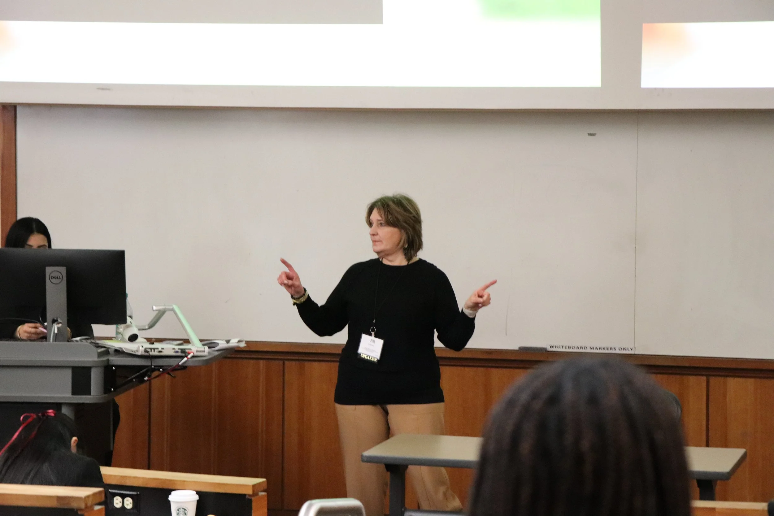Professor standing in front of a classroom whiteboard presenting to students, with students seated and a computer on a cart to her left.