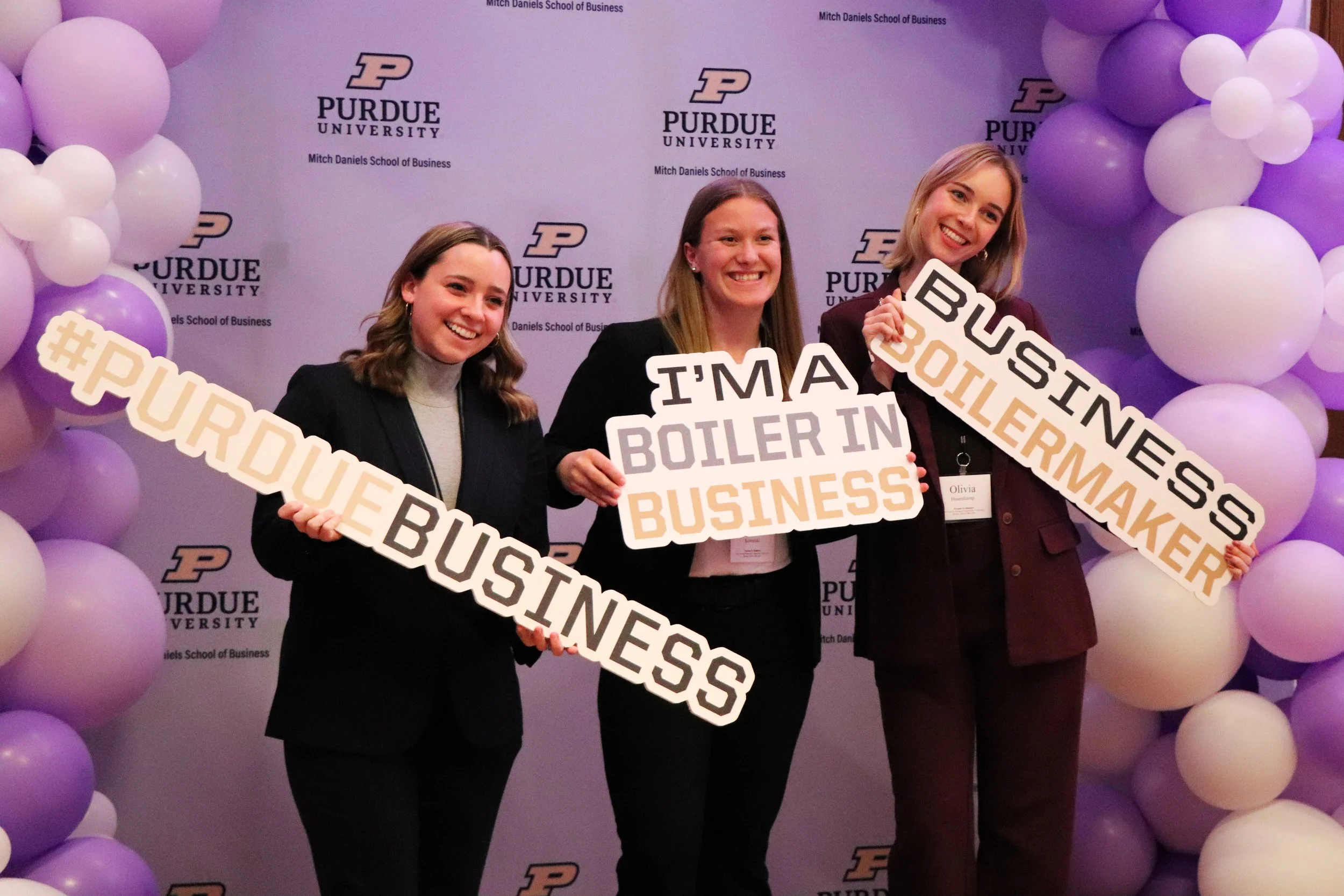 Three women holding Purdue University-themed signs at an event with purple and white balloon decorations and Purdue University logos in the background.