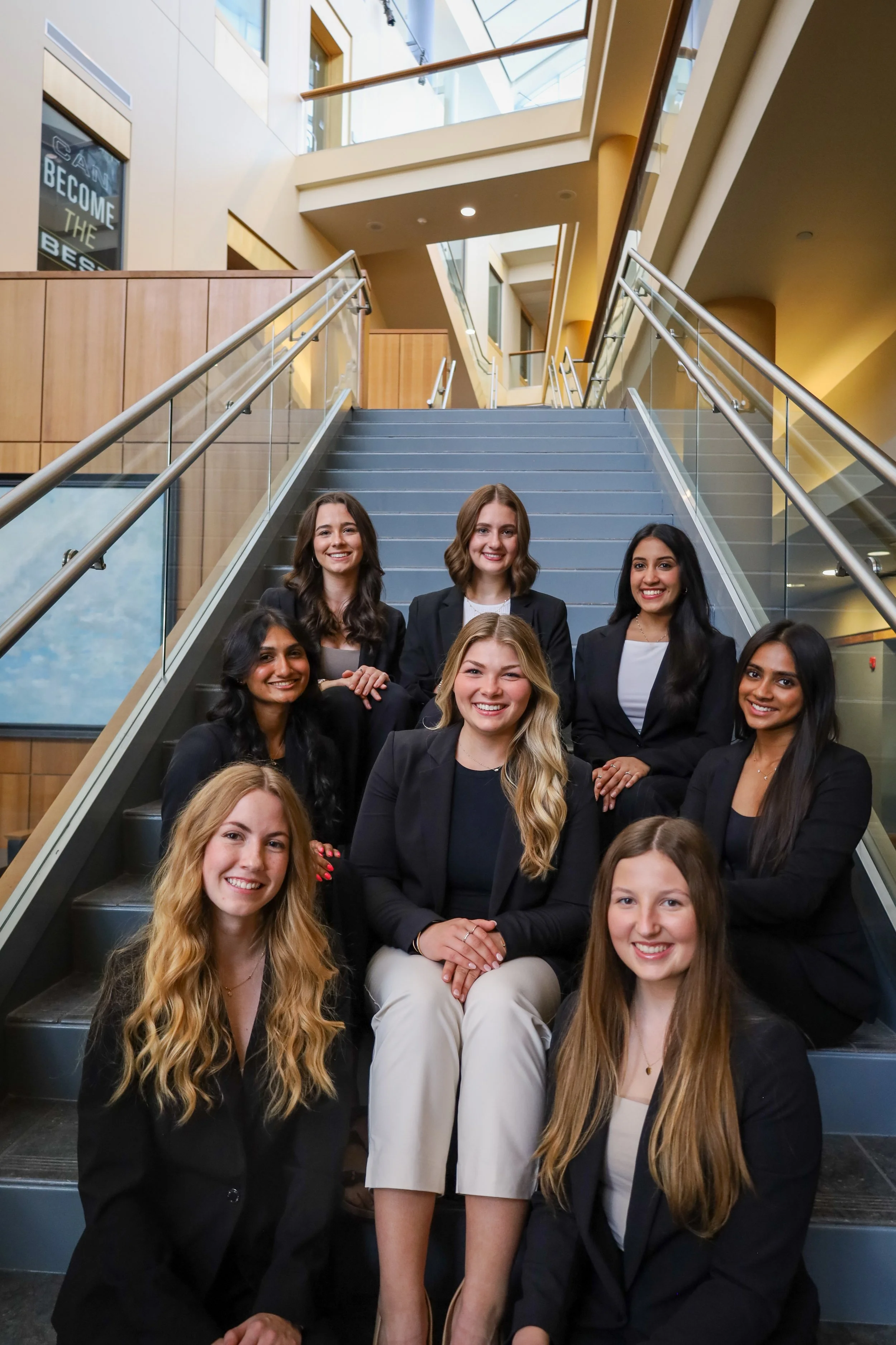 Group of nine diverse women in business attire sitting on stairs in modern office building