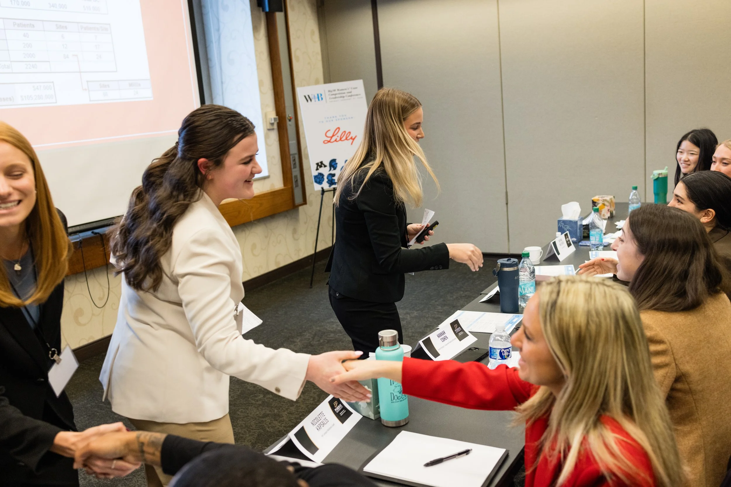 Women at a conference table shaking hands, with some smiling and others listening, in a room with presentation screens and conference materials.