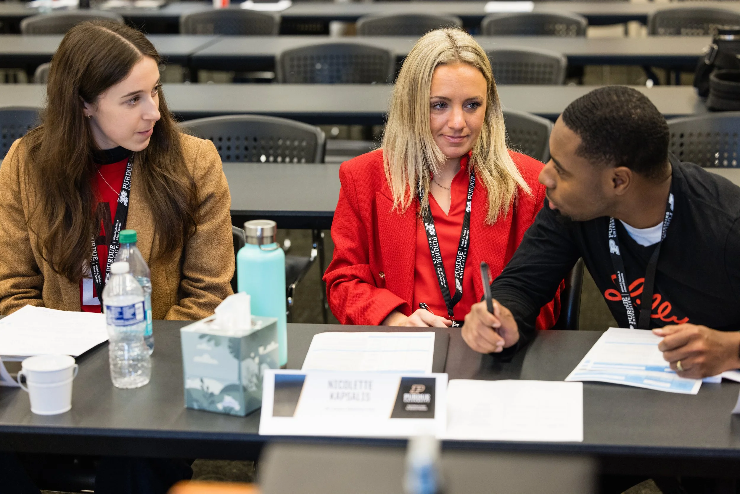 Three young adults sitting at a table, engaged in conversation, with papers, water bottles, and a tissue box on the table, in a conference or classroom setting.