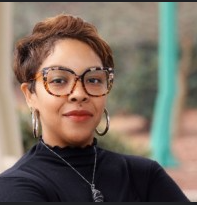 A woman with short curly hair wearing glasses, hoop earrings, and a black top, standing outdoors with trees and playground equipment in the background.